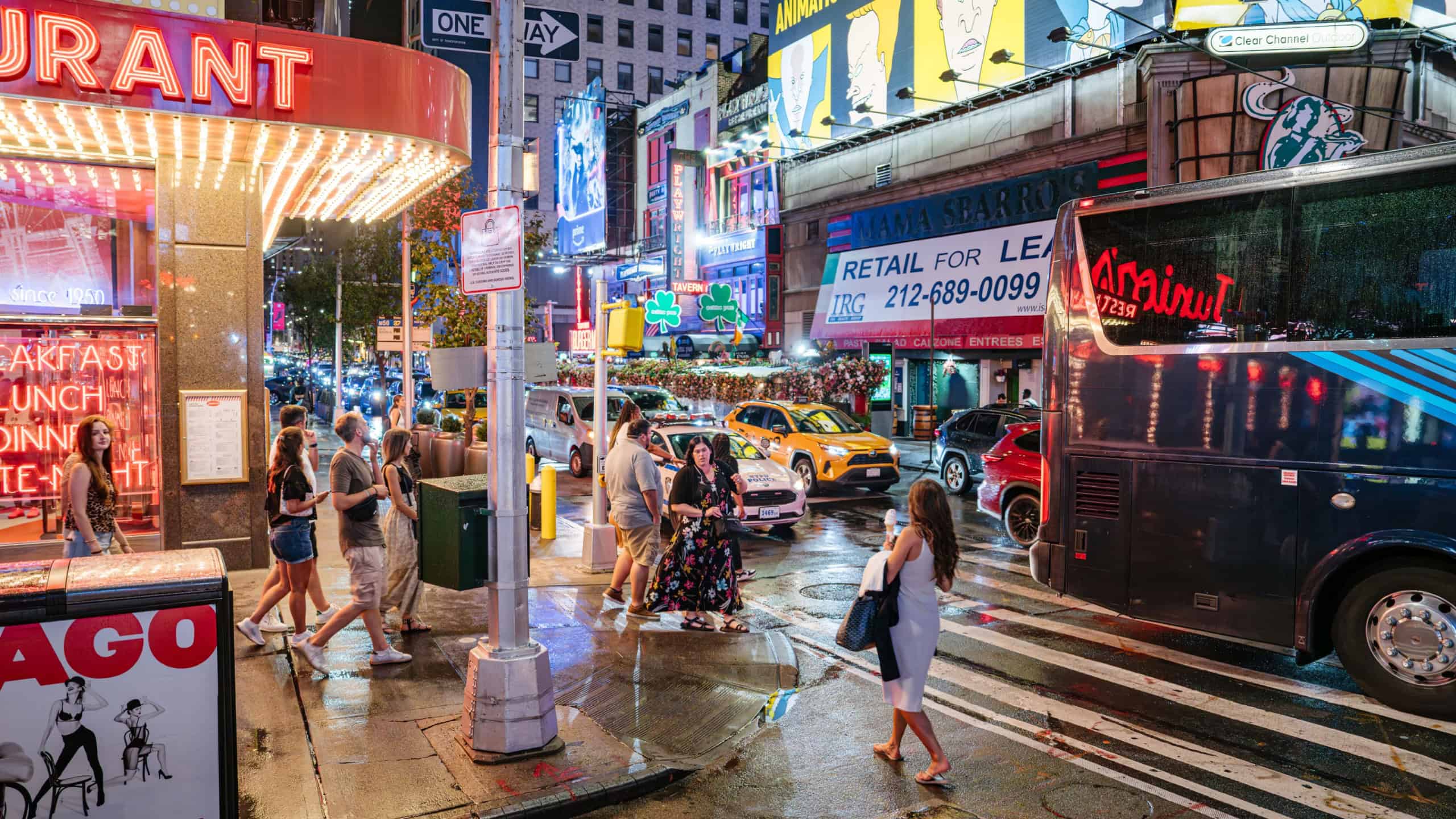 New York City, New York, USA - 07.31.2024: Pedestrians walk along a wet New York City sidewalk near a glowing diner entrance and passing traffic. Neon lights, billboards, and a crowded street reflect