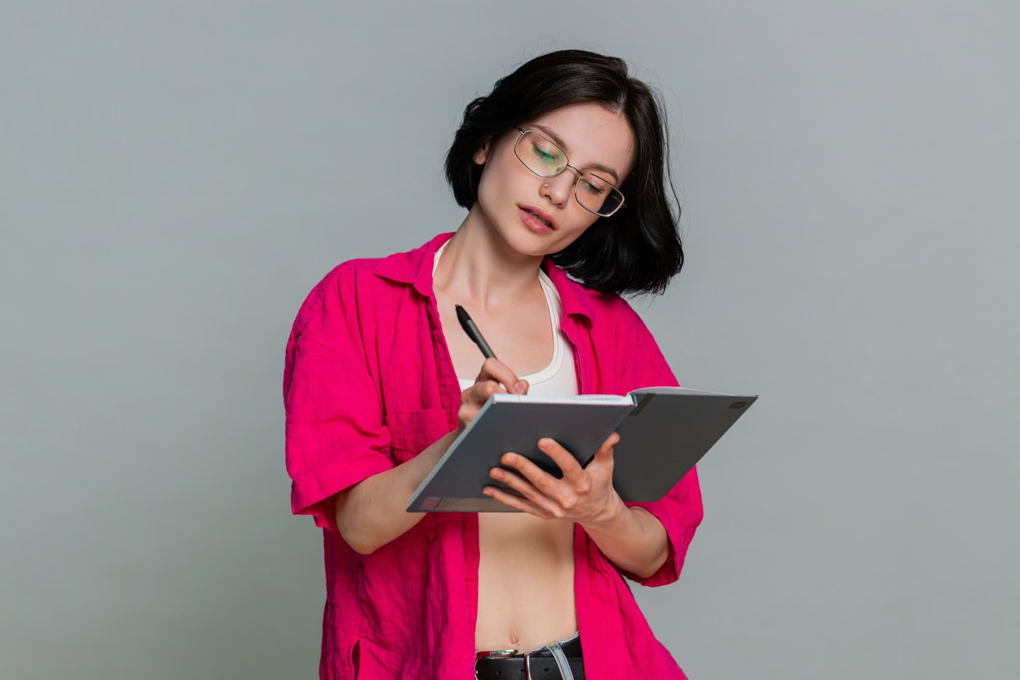 Young woman thoughtfully writes in notebook, carefully creating a checklist and planning upcoming tasks. Girl demonstrates organized mindset and focused planning activity, isolated on gray background.