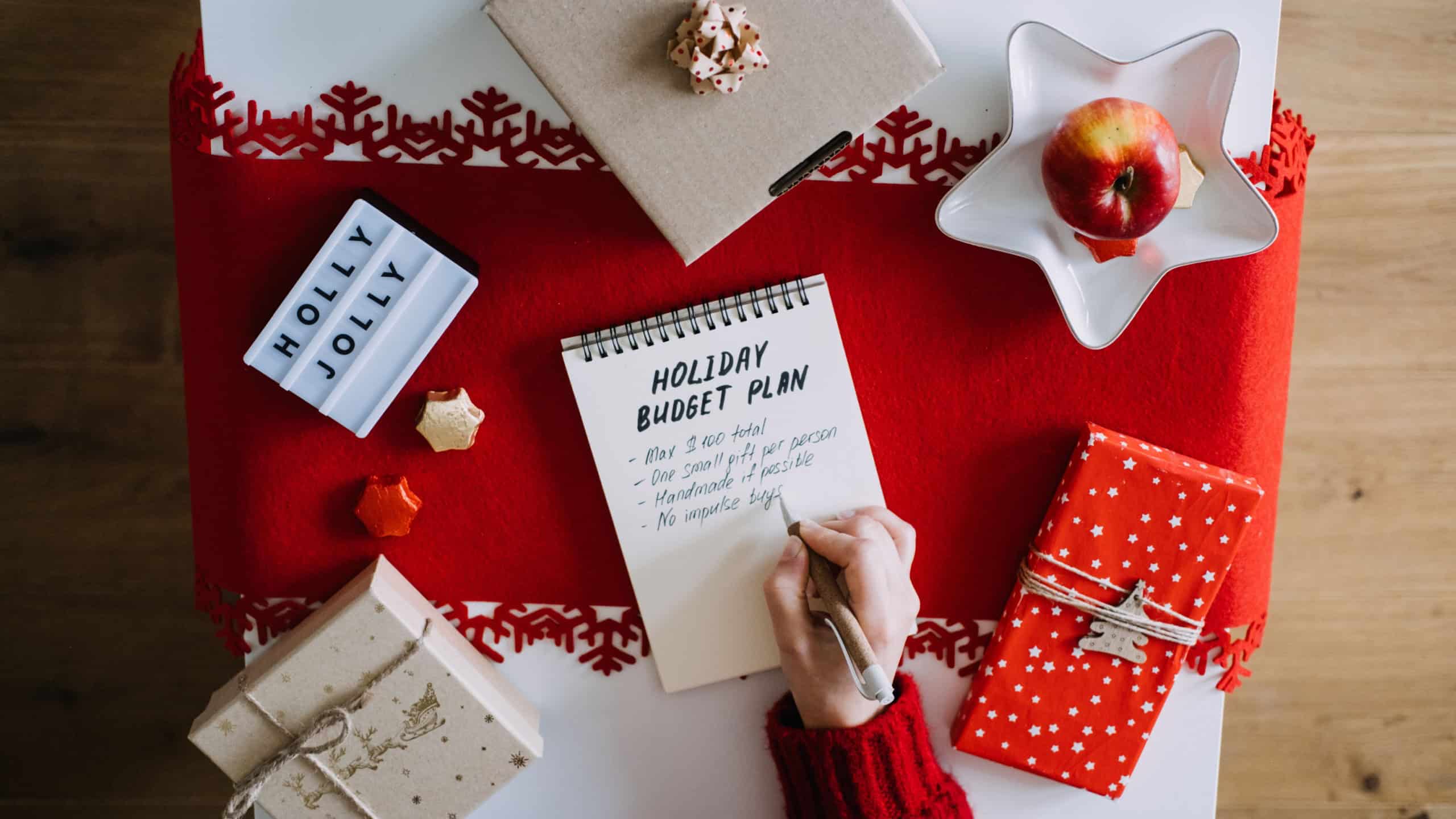 Person writes HOLIDAY BUDGET PLAN in notebook surrounded by wrapped gifts HOLLY JOLLY light box and festive decorations. Holiday budgeting, minimalist gifting, financial wellness, Christmas planning.