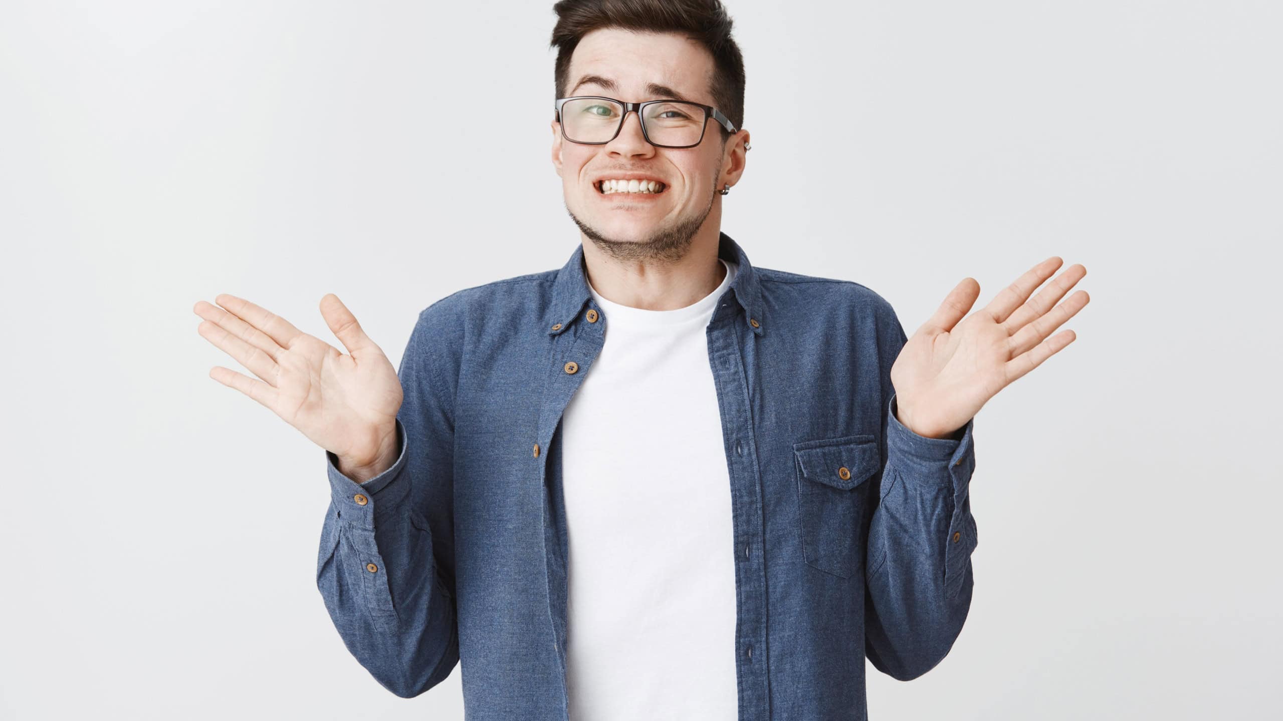 Sorry I messed up. Guilty young and handsome male in glasses and blue shirt raising hands near shoulders, shrugging making awkard smile as apologizing for making mistake over white background