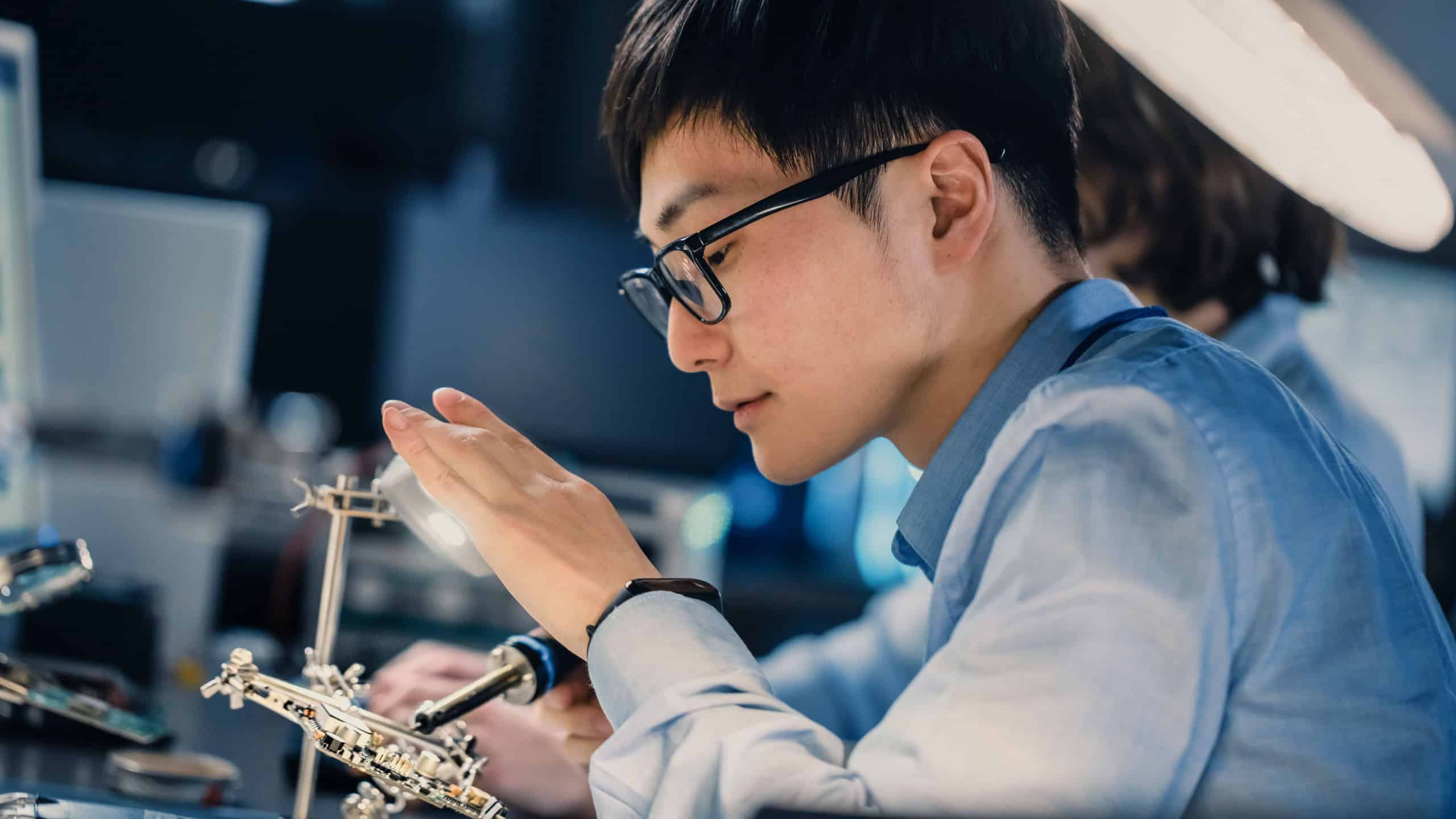 Close Up of a Professional Japanese Electronics Development Engineer in Blue Shirt Soldering a Circuit Board in a High Tech Research Laboratory with Modern Computer Equipment.