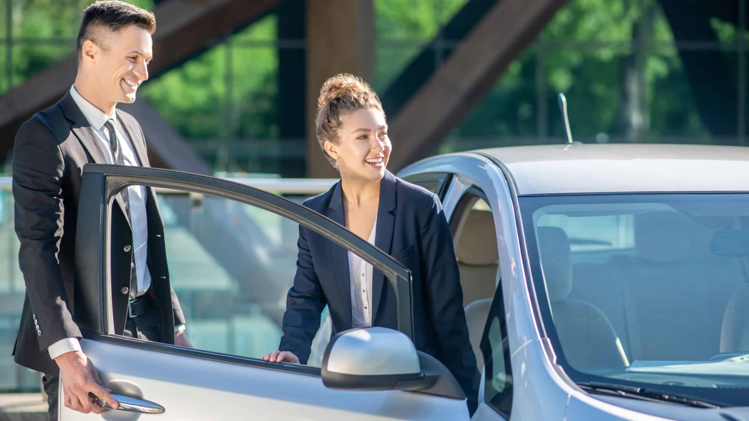 Lucky day. Joyful woman and successful man in formal business suits are standing smiling near the open car door.