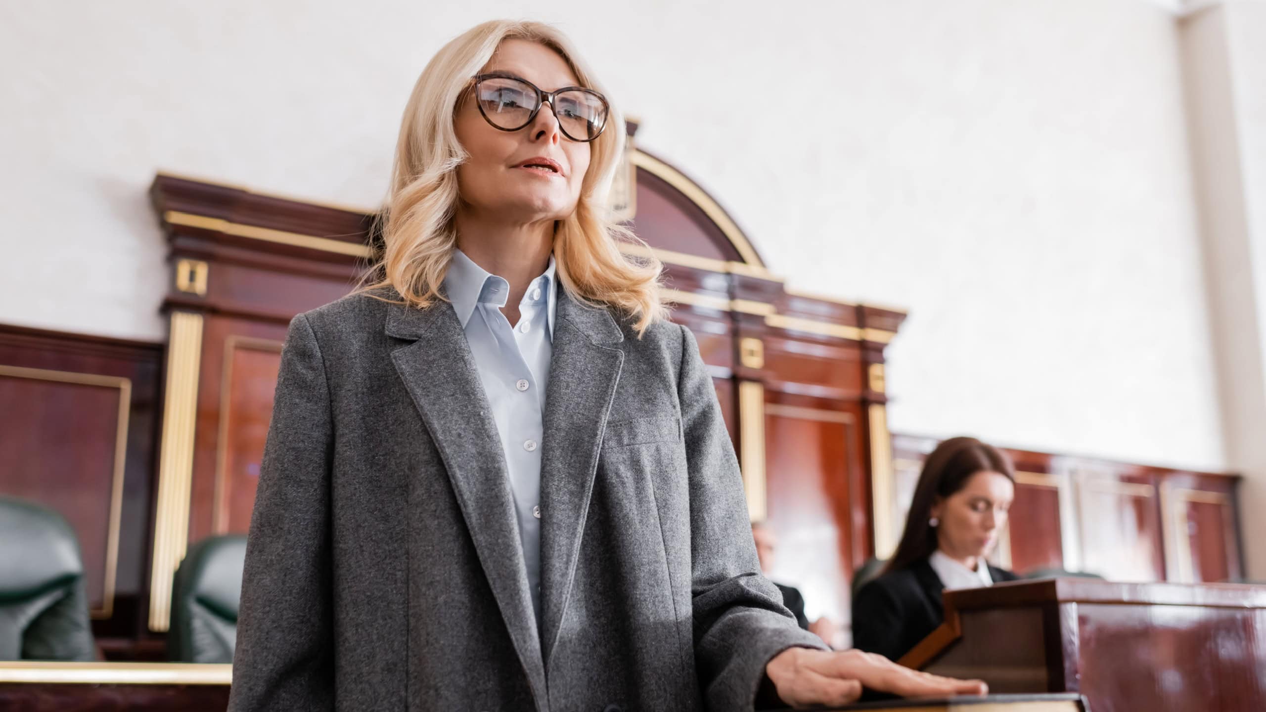 woman in eyeglasses giving swear on bible in court near prosecutor on blurred background