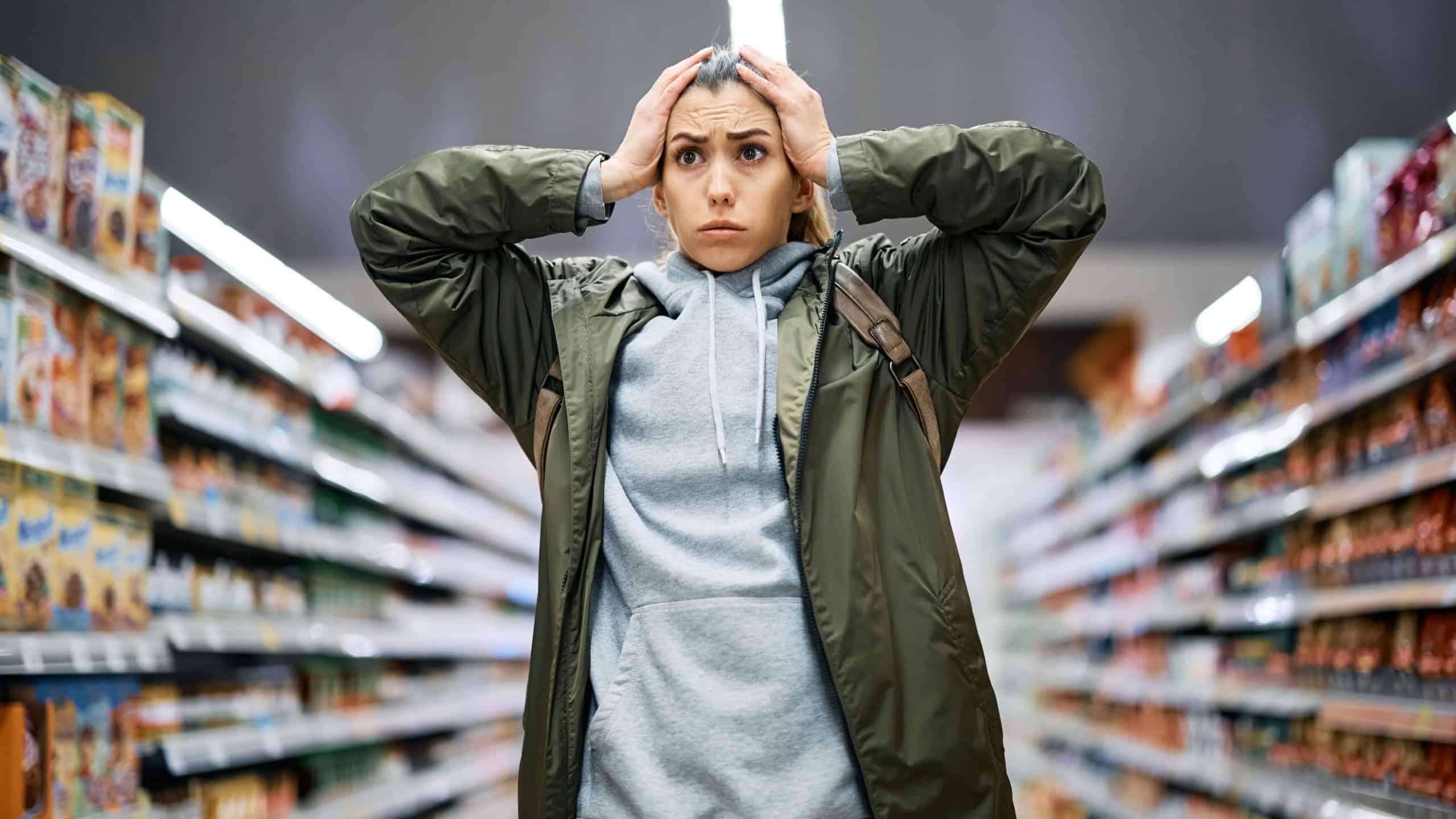 Young woman feeling shocked about rising food prices while standing among the aisle with groceries in supermarket.