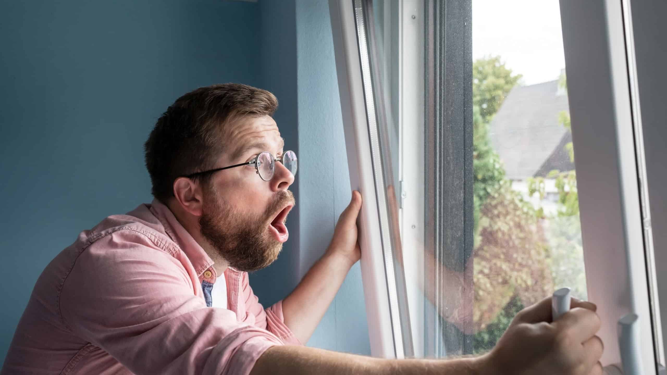 Shocked, frightened man looks out the window and screams excitedly, standing against the background of the blue wall in the room.