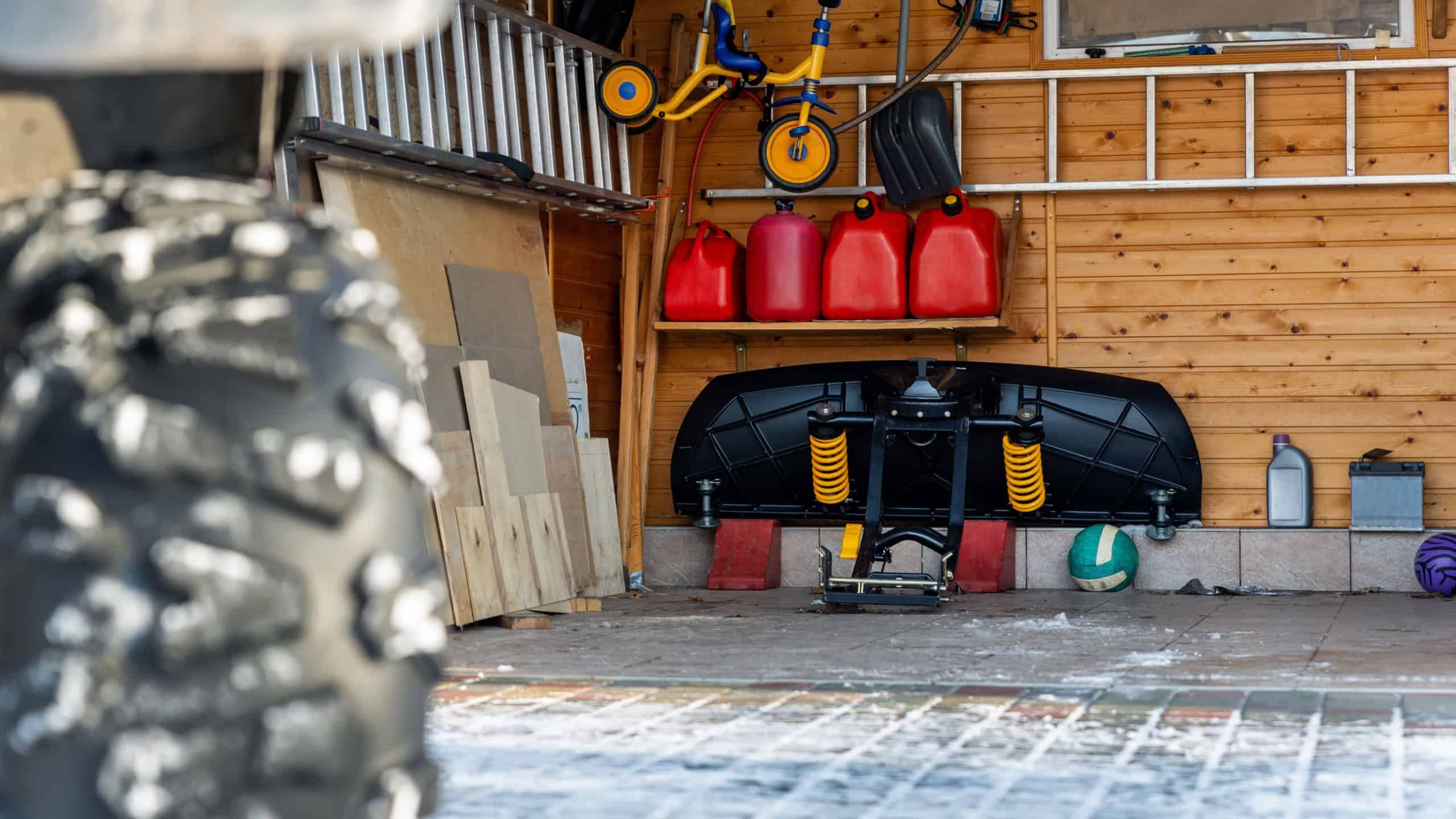 Back POV close-up detail view of quad bike offroad vehicle parked front house garage open door on sunny snowy cold winter day. Scoop snow removal tool equipment at home. Organized stuff storage shed