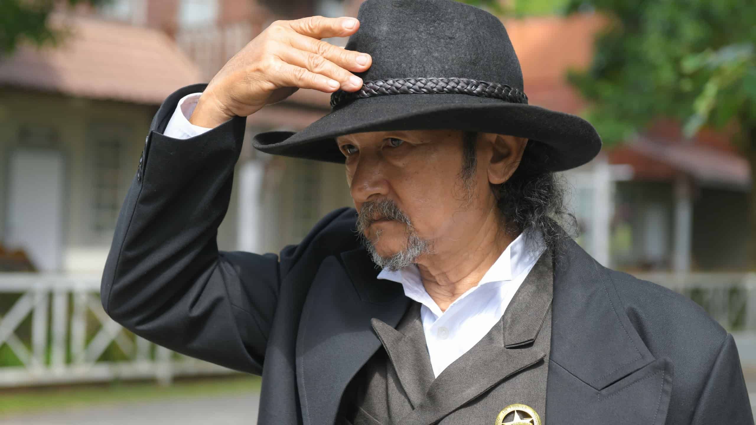 The sheriff dressed in a uniform with the Sheriff's star badge, he is raising his hand to take off his leather brimmed hat.