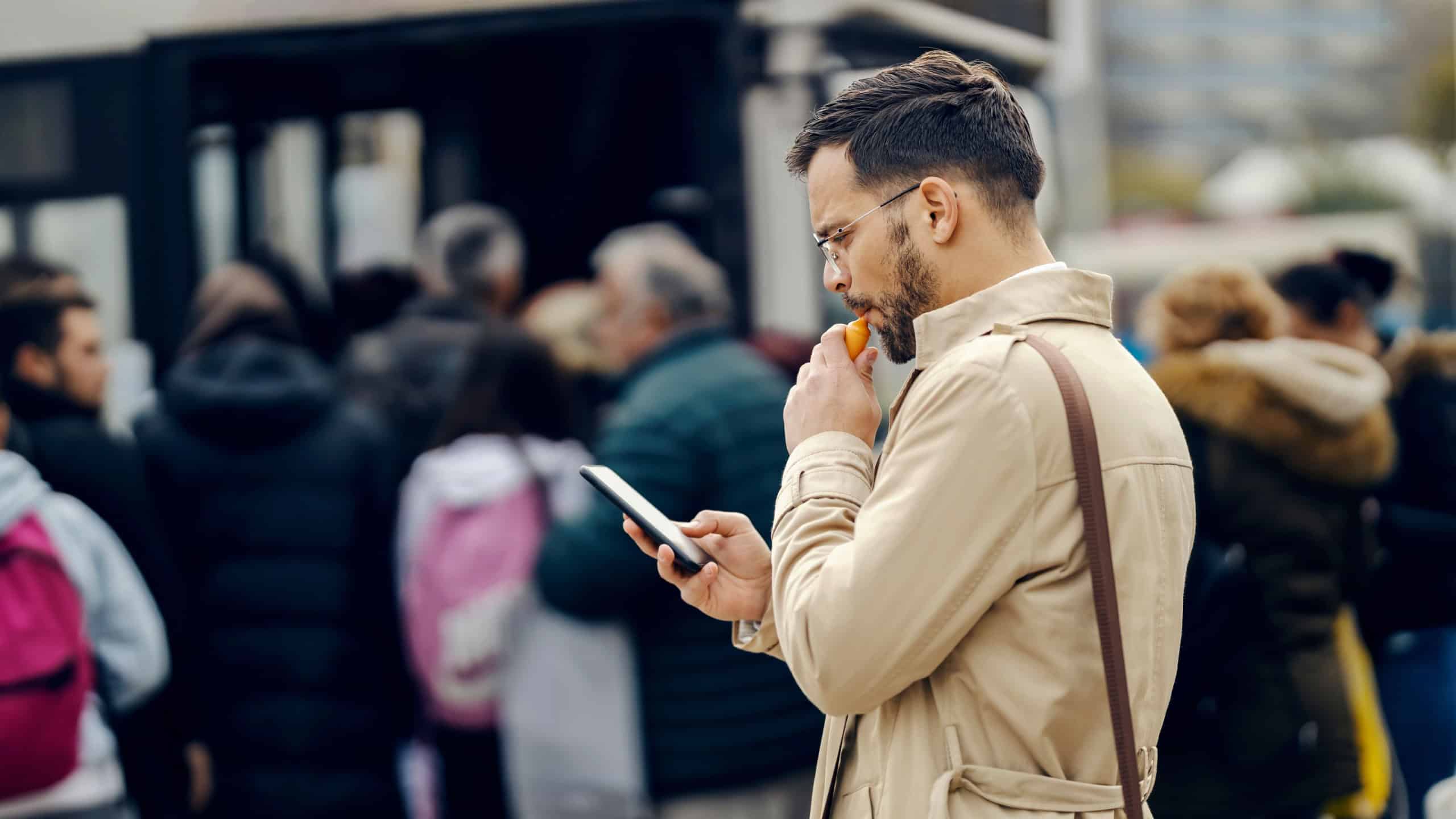A trendy businessman is smoking vape and looking at the phone while standing downtown in crowd.
