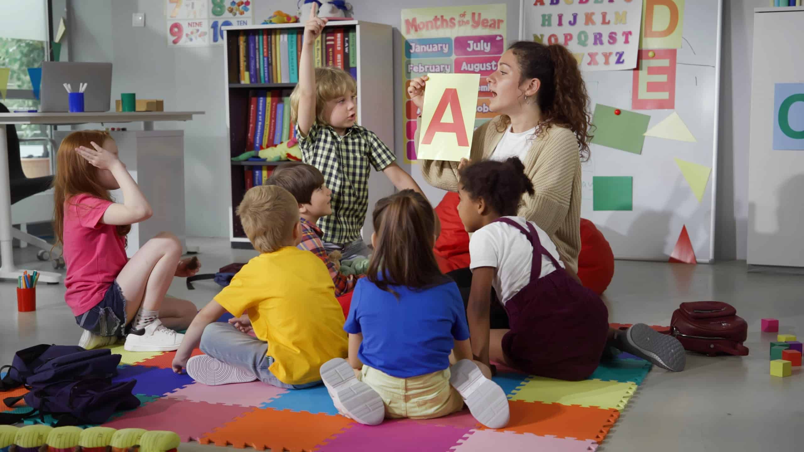 Female teacher hold flashcard with letter a teaching preschool kids alphabet. Diverse pupils of primary school sit on floor with teacher having alphabet lesson