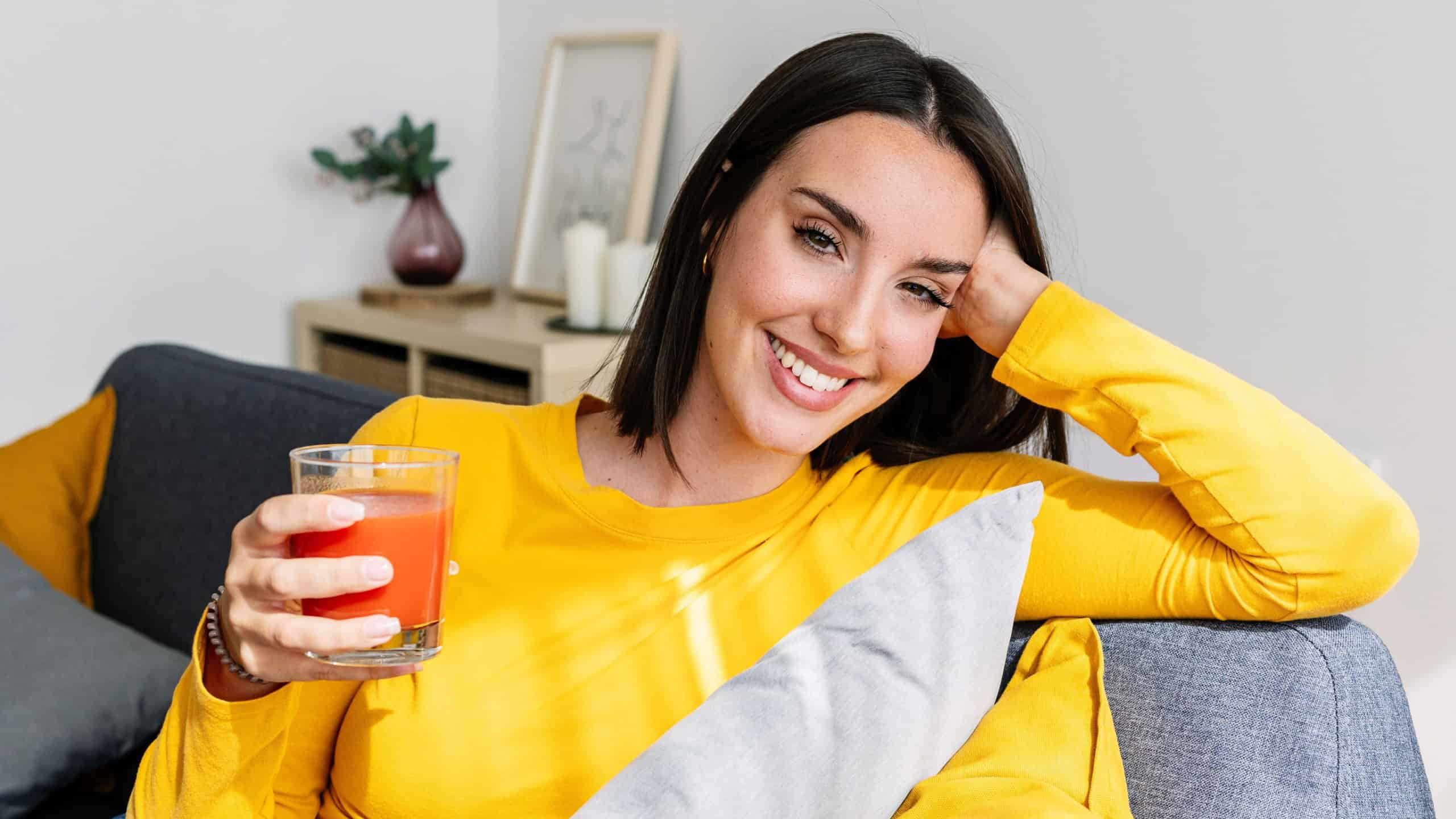 Smiling portrait of young beautiful woman drinking tomato juice relaxing on couch at apartment. Healthy lifestyle and well-being concept.