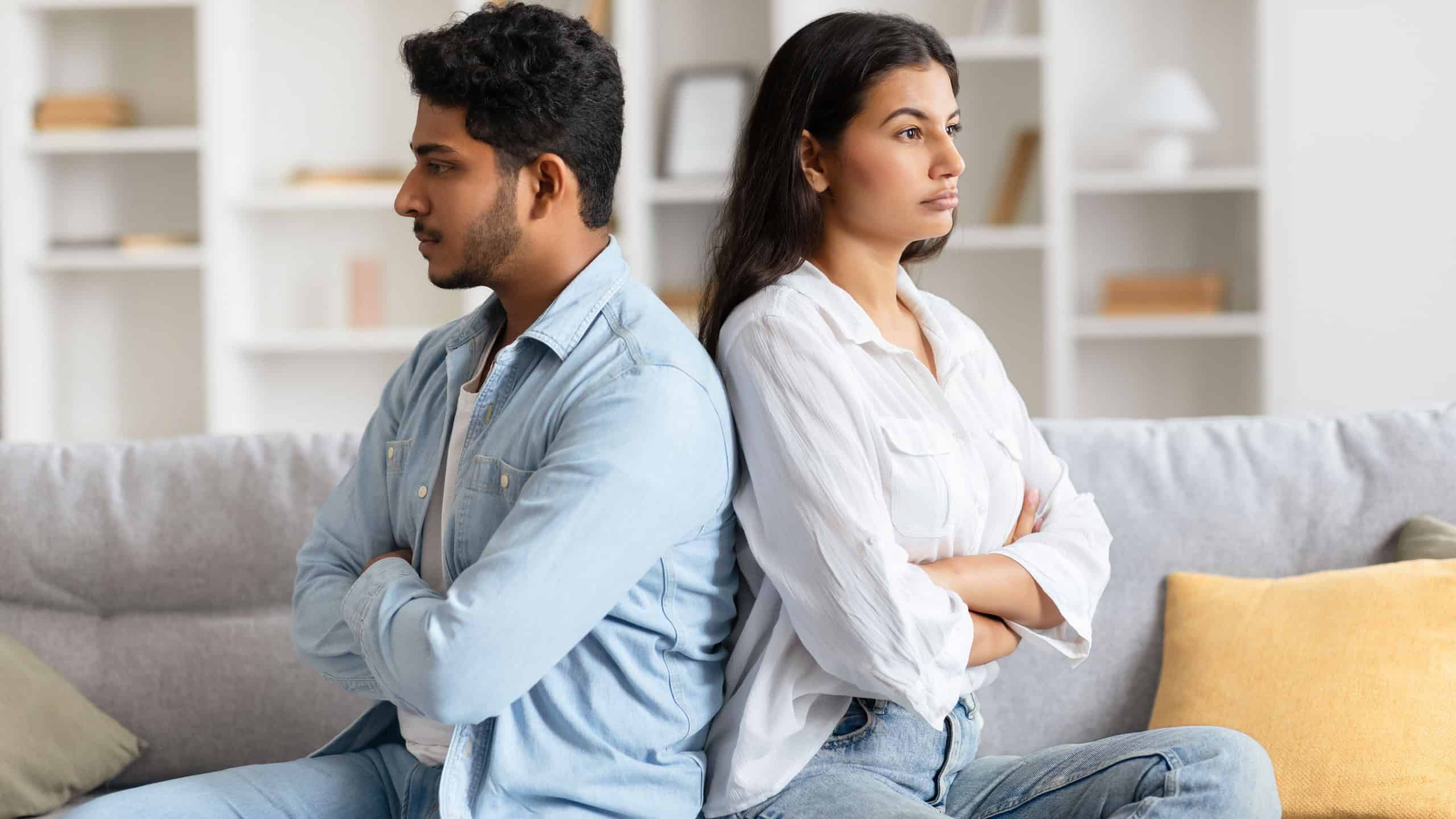 Young indian couple sits back-to-back on sofa, surrounded by a tense atmosphere, visibly upset and offended following a heated quarrel in their homey living space