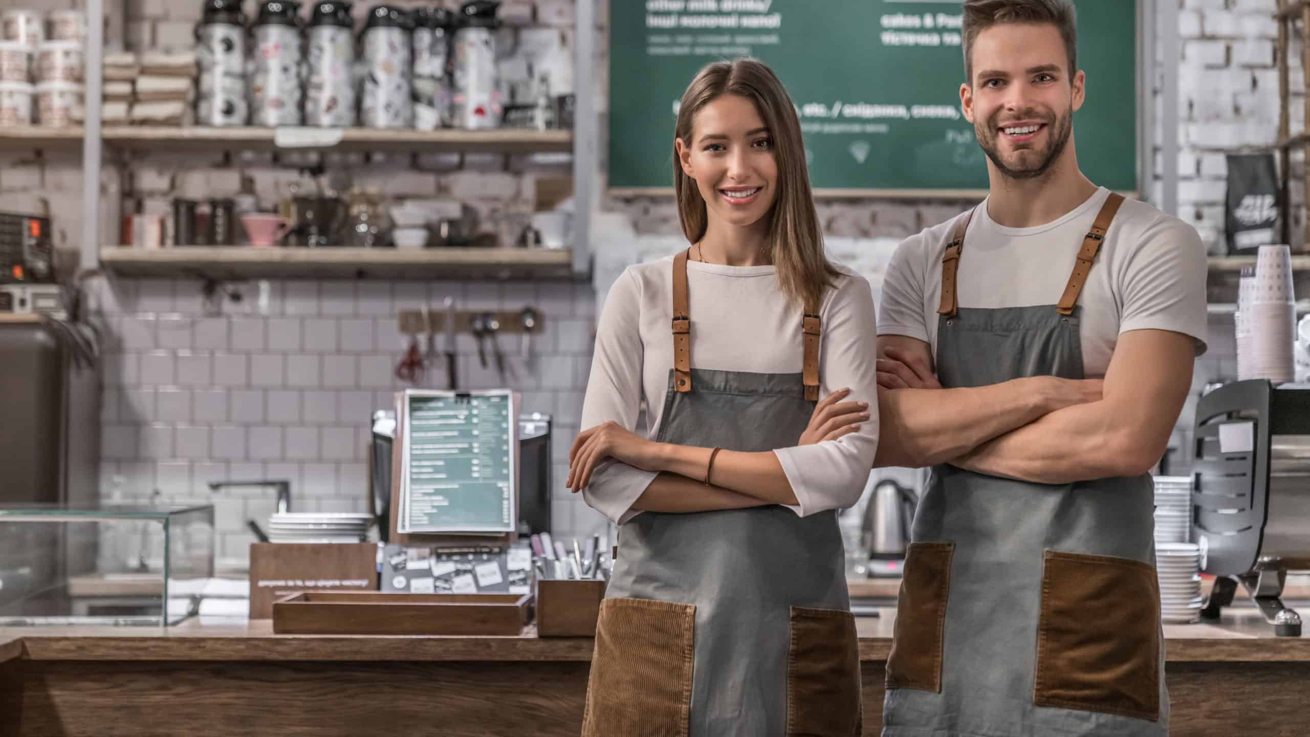 Portrait of successful business coffee shop owners indoors. Photo of young caucasian waiter and waitress standing in uniform with arms crossed and smiling at camera with counter on background
