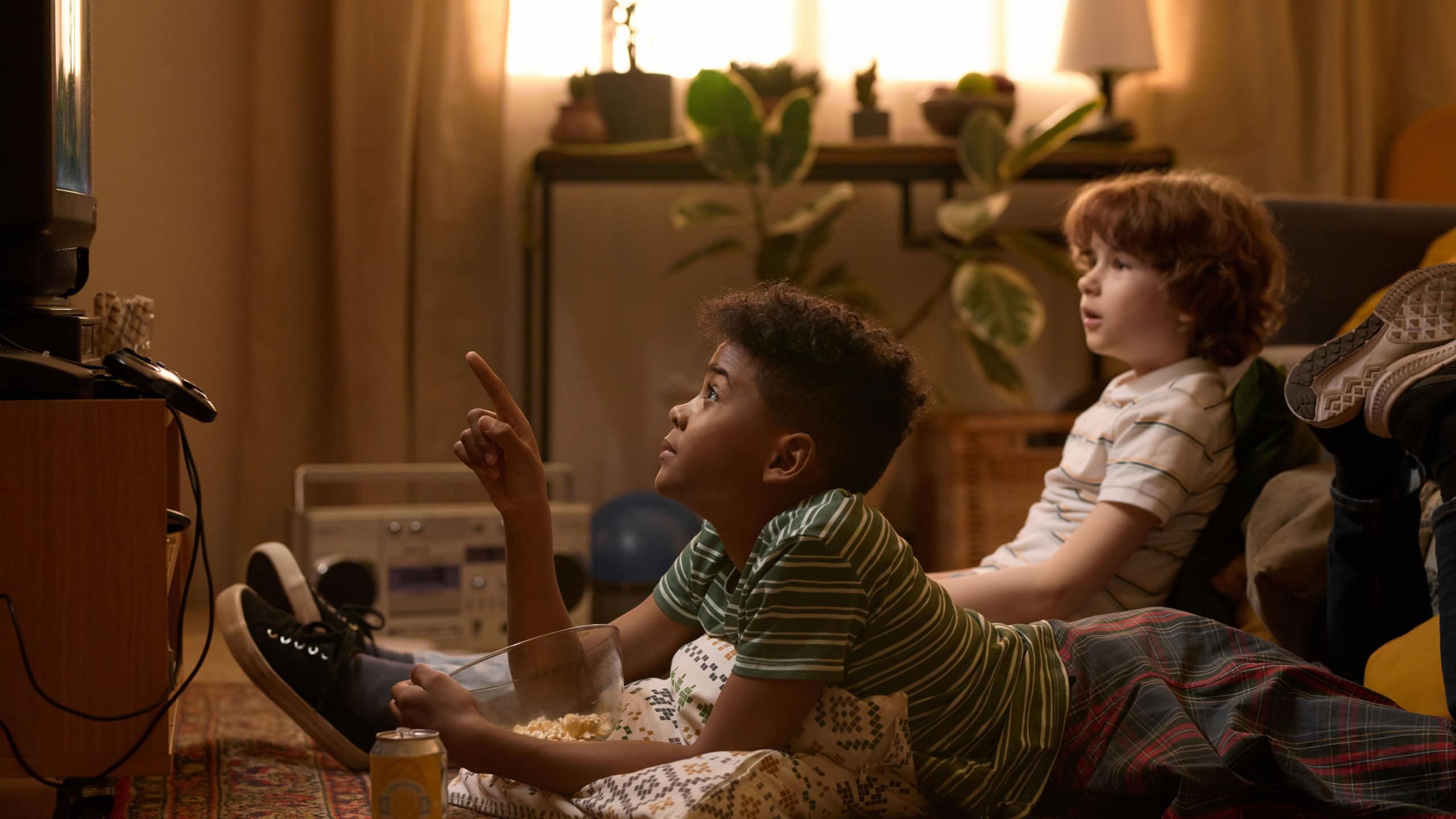Two multiethnic schoolkids lying on floor, one pointing at TV while watching exciting movie in cozy retro-styled living room with relaxed atmosphere and snacks like popcorn and soda