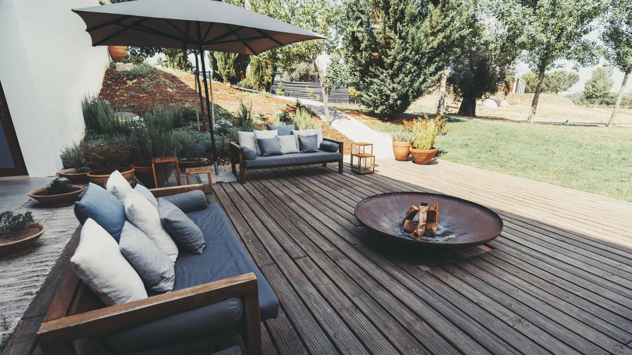 Outdoor patio area with wooden deck, cushioned sofas, and a fire pit. An umbrella provides shade, and the garden is landscaped with various plants and trees, creating a cozy ambiance