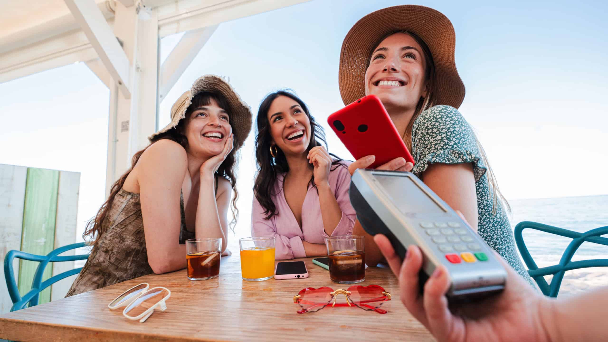 Happy young group of only women paying the bill with a nfc cellphone in cocktail beach bar. Female holding a smart phone and giving a payment transaction to the cashier on vacations with her friends