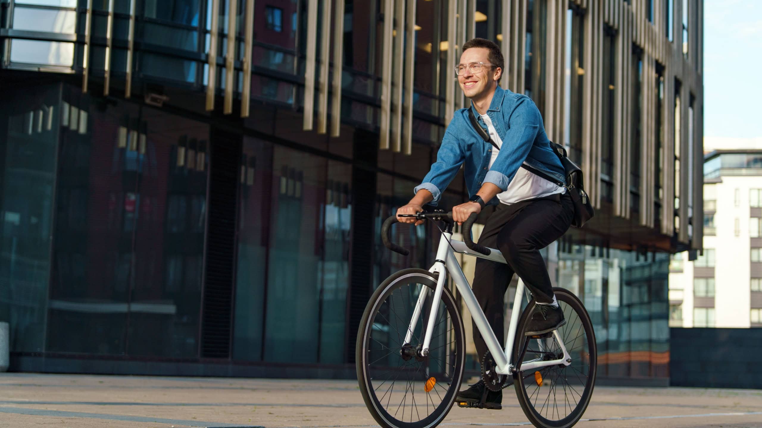 Smiling man in a casual denim jacket riding a white bicycle through a modern cityscape during the day.