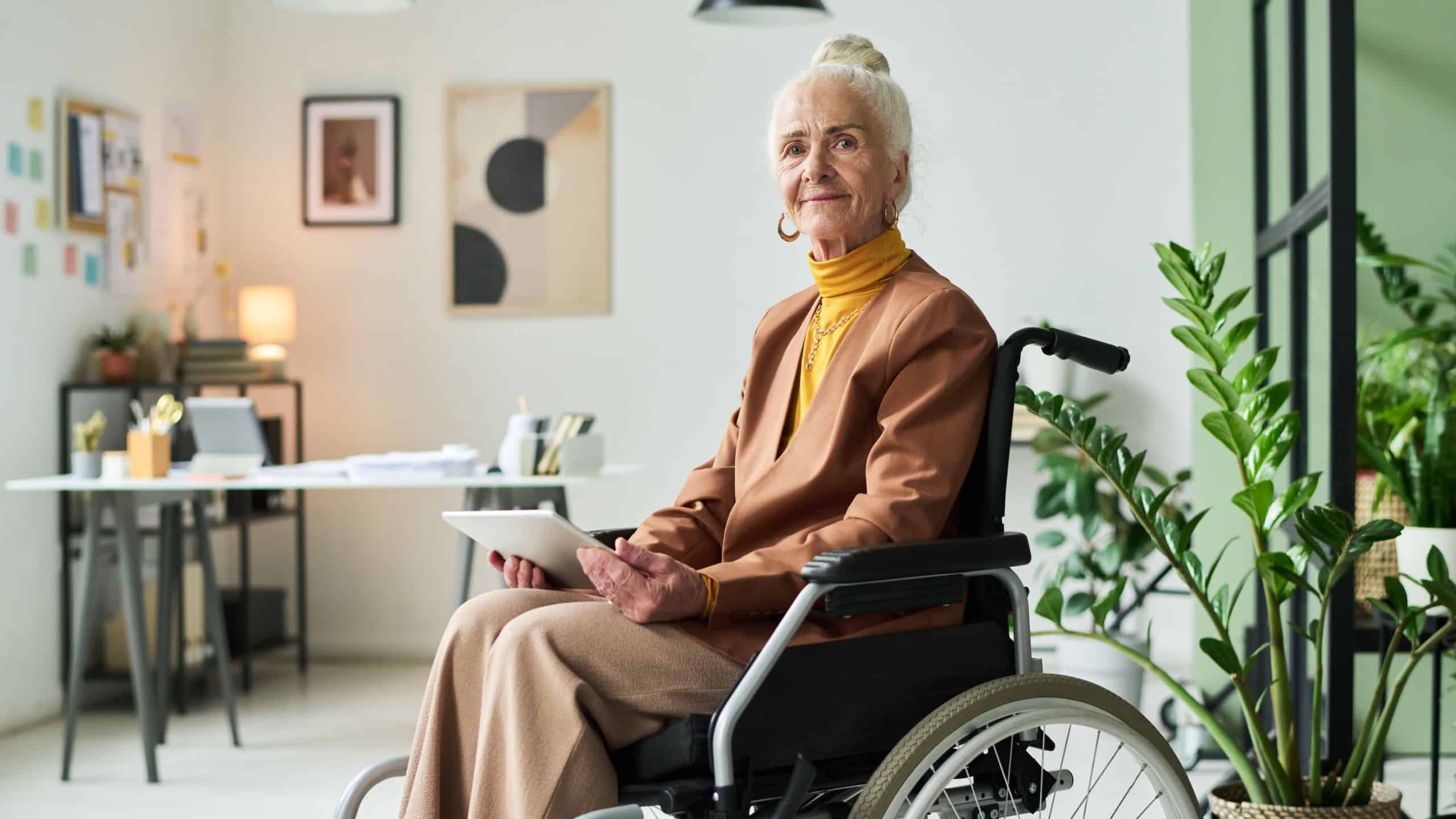 Elderly woman sitting in wheelchair in modern office holding tablet and looking at camera, with plants and office supplies in background, creating a professional atmosphere