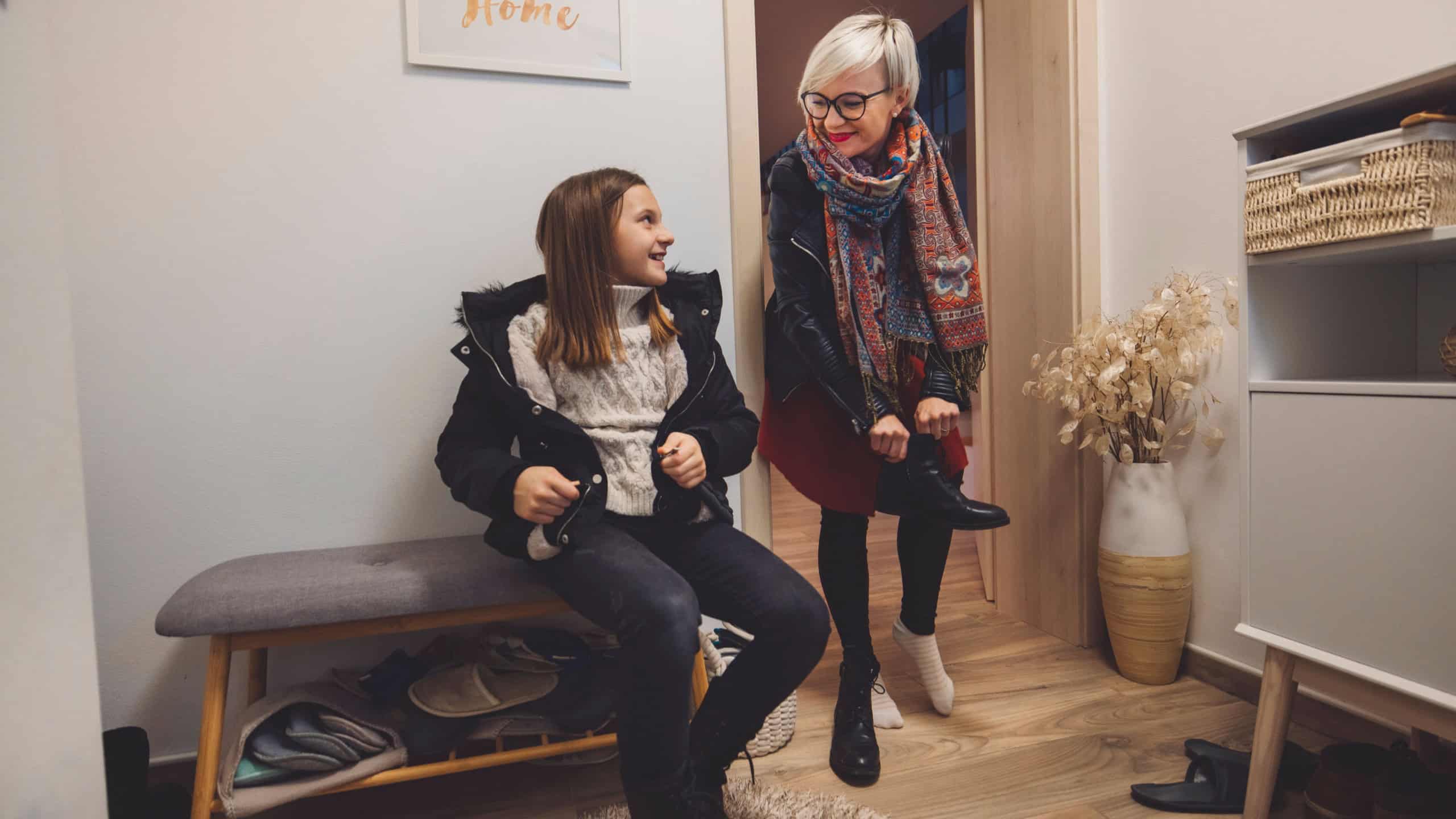 A cozy home entrance scene featuring a young girl sitting on a bench, putting on her shoes, while an older woman stands nearby, smiling and engaging with her. The decor includes a wall art piece that