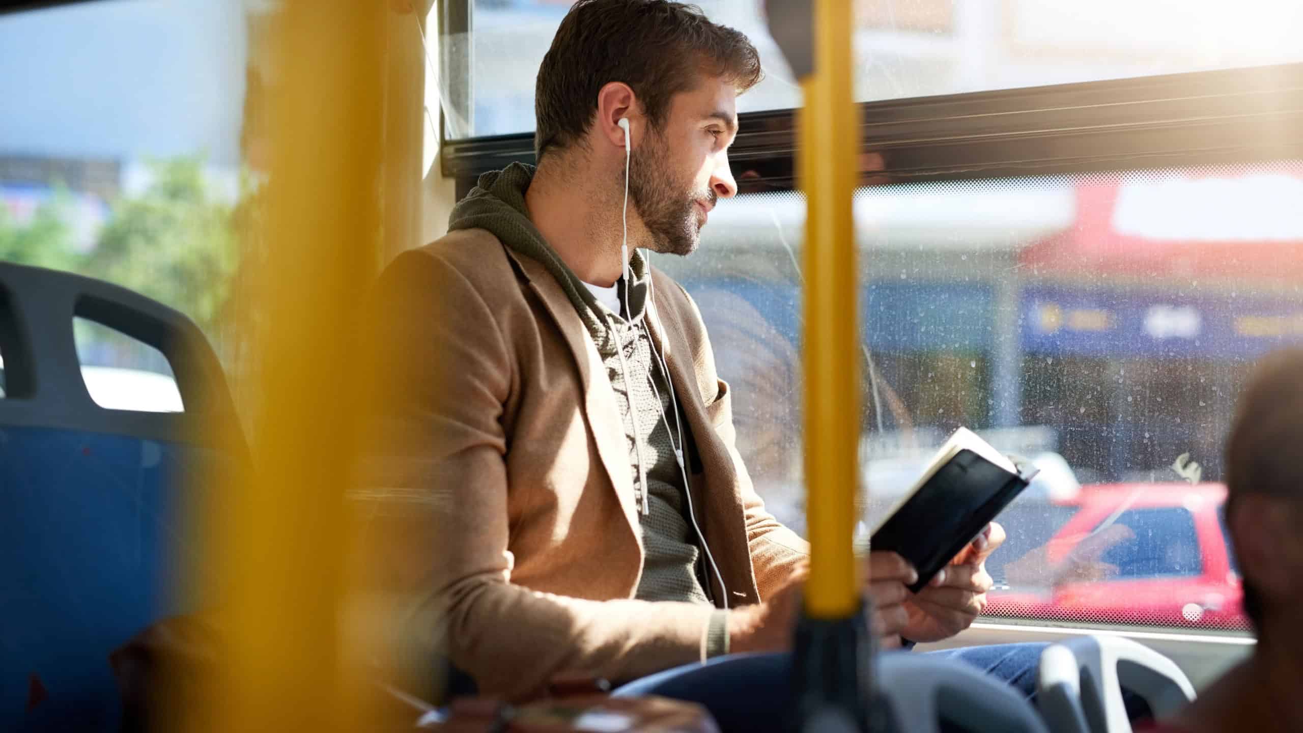 Bus, travel and man by window with earphones for journey, commute and passenger in city. Public transport, station and person on vehicle with book for reading on trip in urban town, road and metro