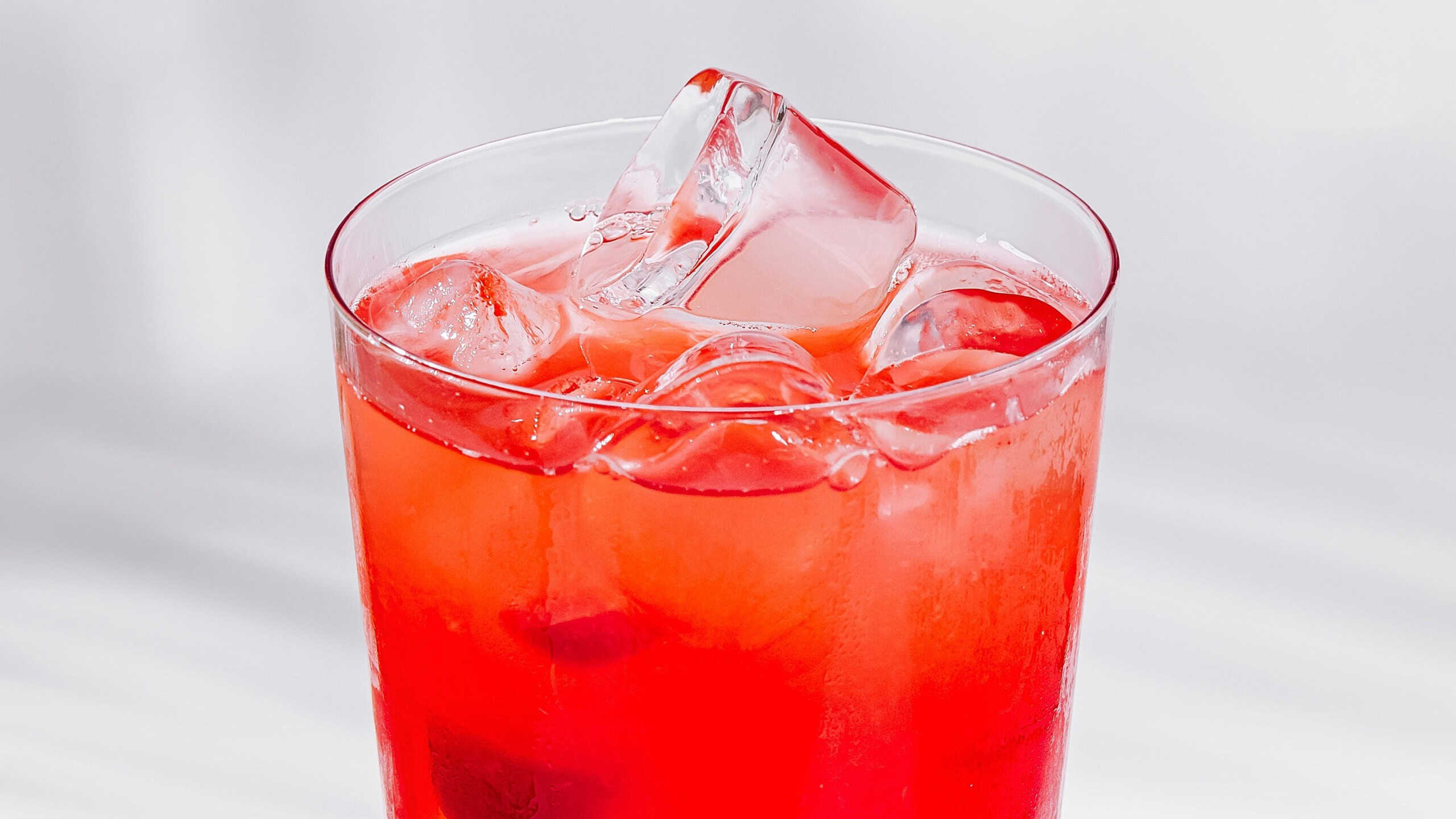 Red cocktail with ice cubes standing on white cube with palm leaf shadow on white background