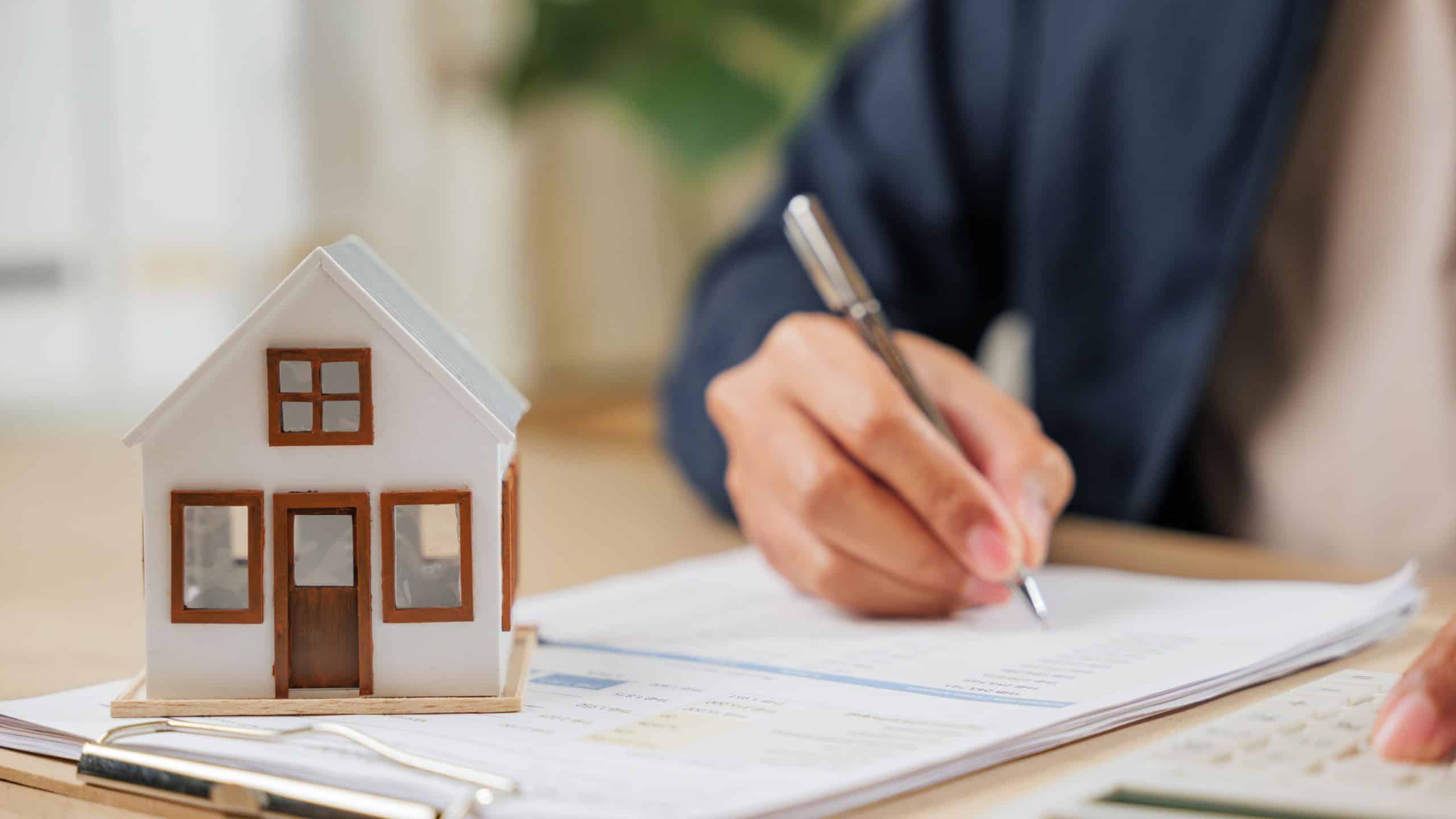 A man writes a signature on house insurance documents, using a calculator for tax and property details. Counting payments and rent by model house, he finalizes the price and investment agreement.