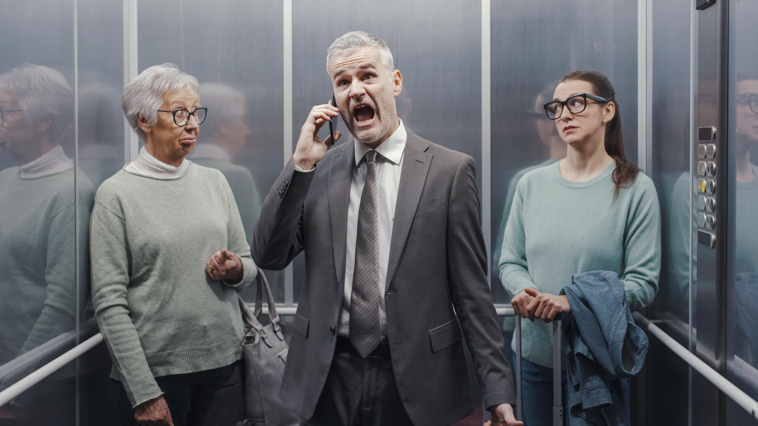 Stressed angry businessman shouting into his smartphone in the elevator, two women are annoyed and looking at each other