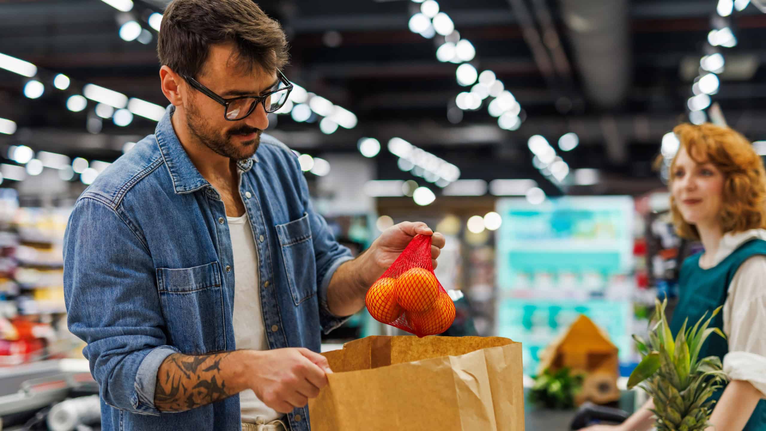 Young man putting oranges into a paper bag at the checkout of a modern supermarket while a cashier waits in the background