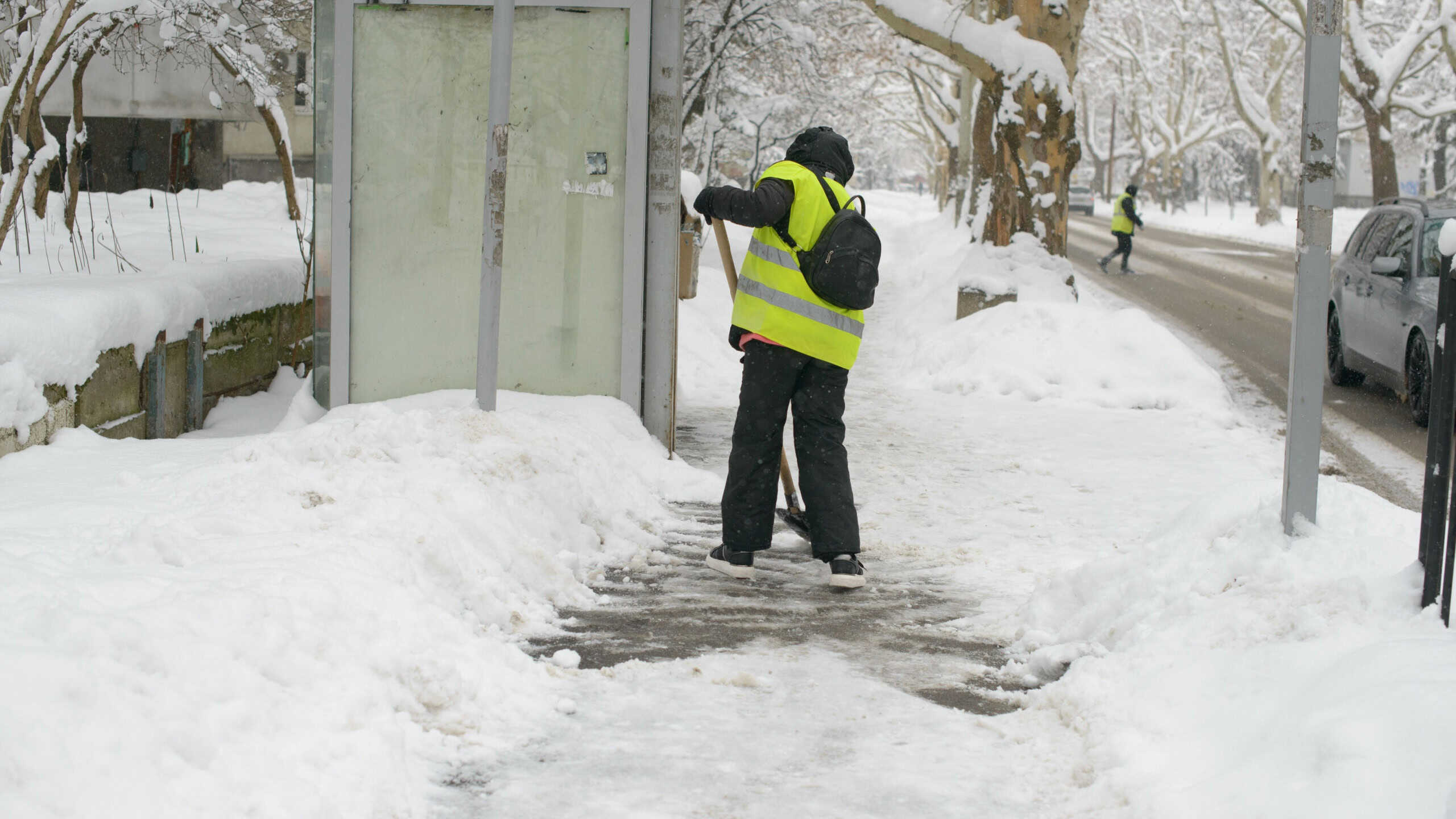 A city worker in a reflective vest shovels snow from the sidewalk beside a bus stop, ensuring pedestrian safety and clear pathways in winter conditions