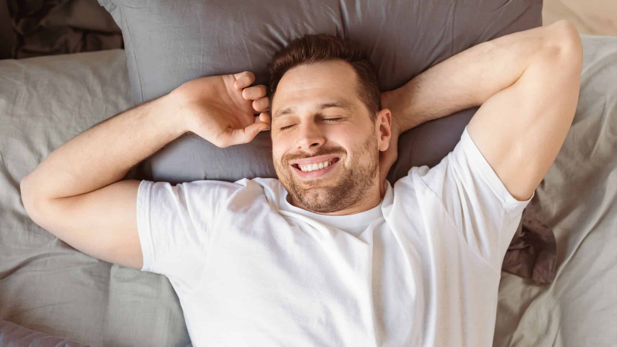 Good Morning. Joyful Guy Waking Up Yawning And Stretching Hands Lying In Comfortable Bed In Bedroom At Home, Smiling With Eyes Closed. Healthy Male Sleep. View From Above, Panorama