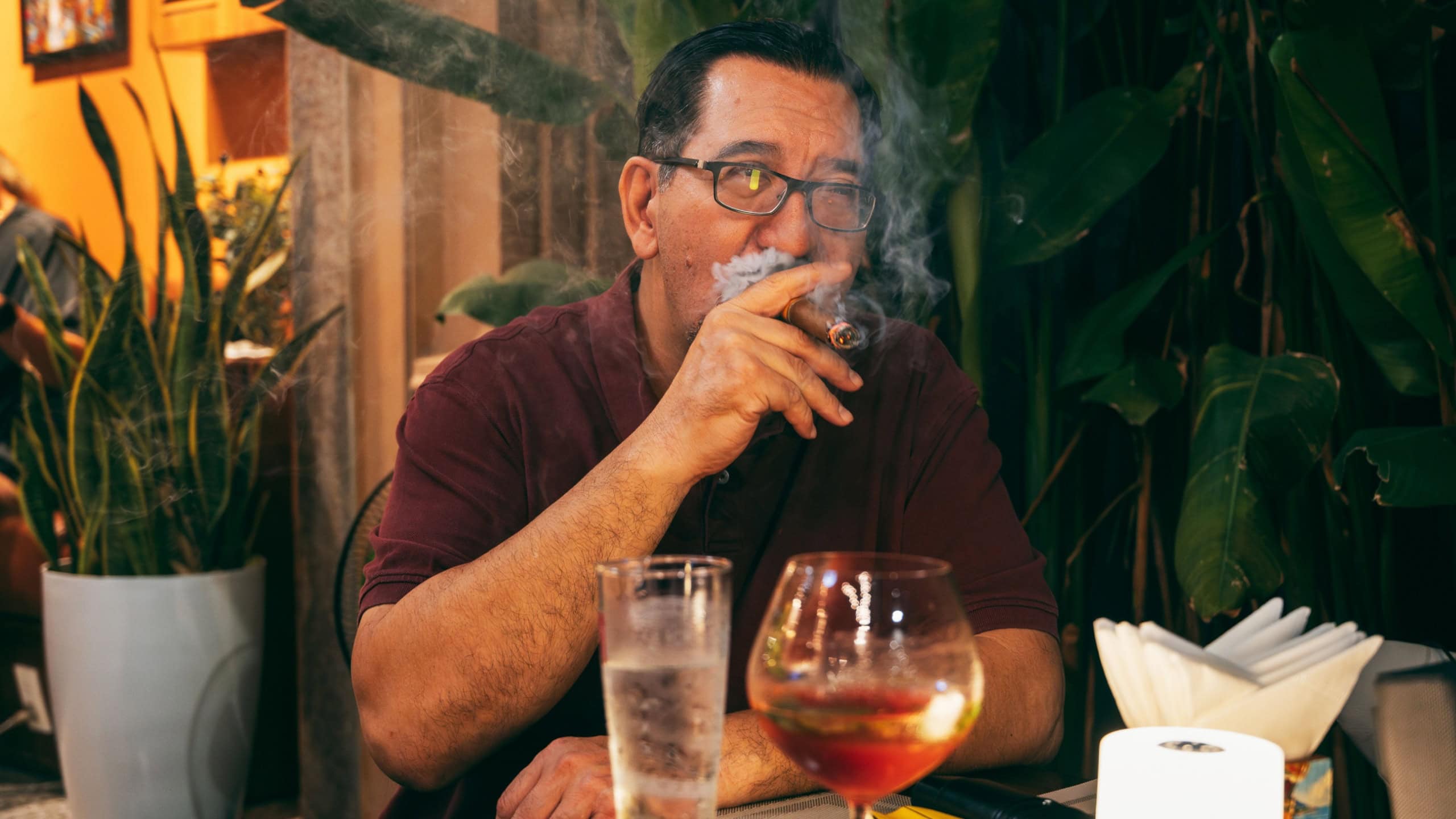 Happy confident successful adult Caucasian man smokes a cigar at restaurant table