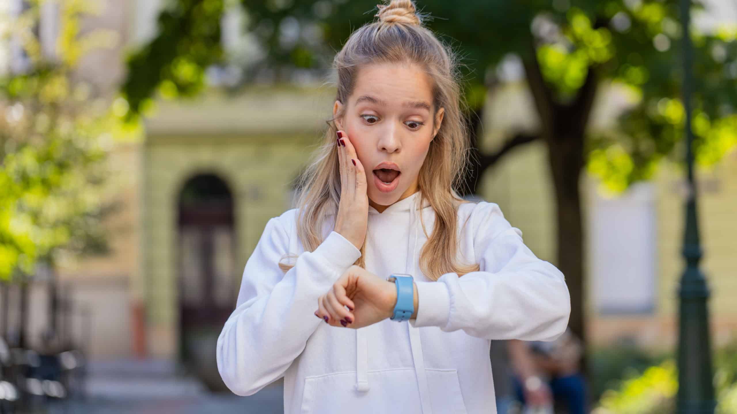 Young woman worrying to be punctual, with anxiety checking time on watch, running late to transport, being in delay, deadline outdoors. Girl teenager tourist walking in city street. Town lifestyles.