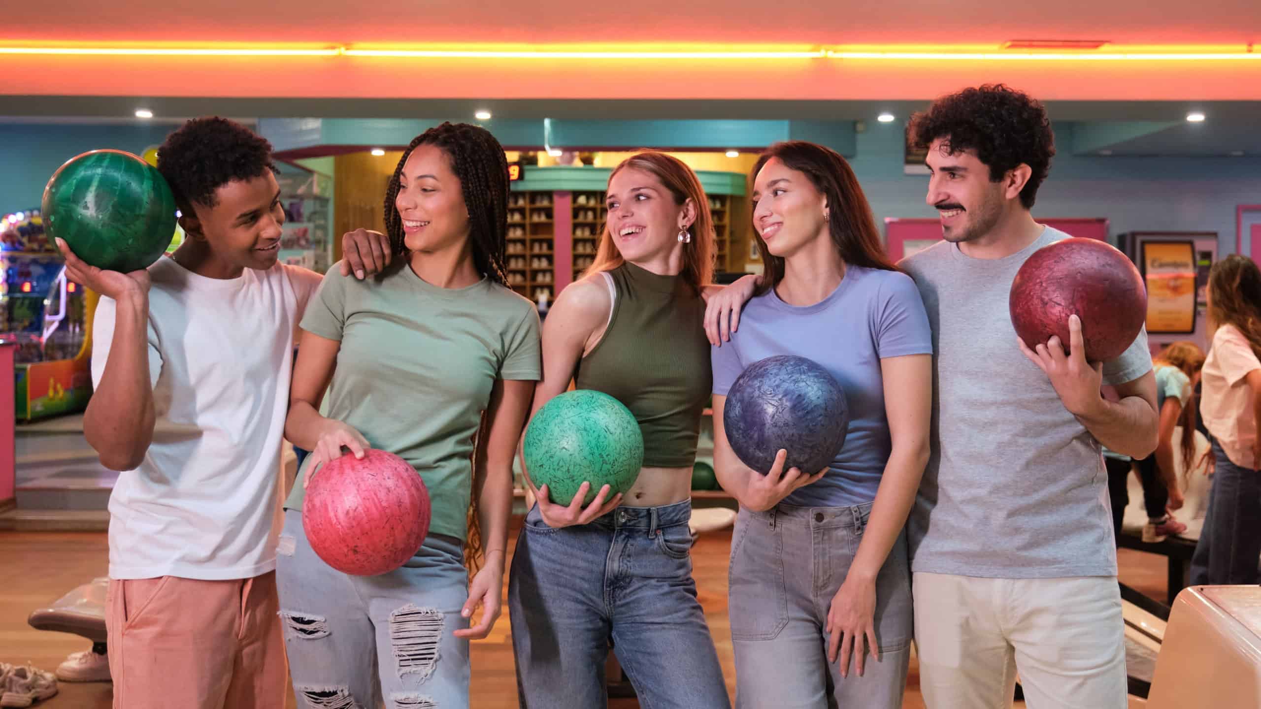 Five friends are holding bowling balls and smiling in a bowling alley, enjoying their leisure time together