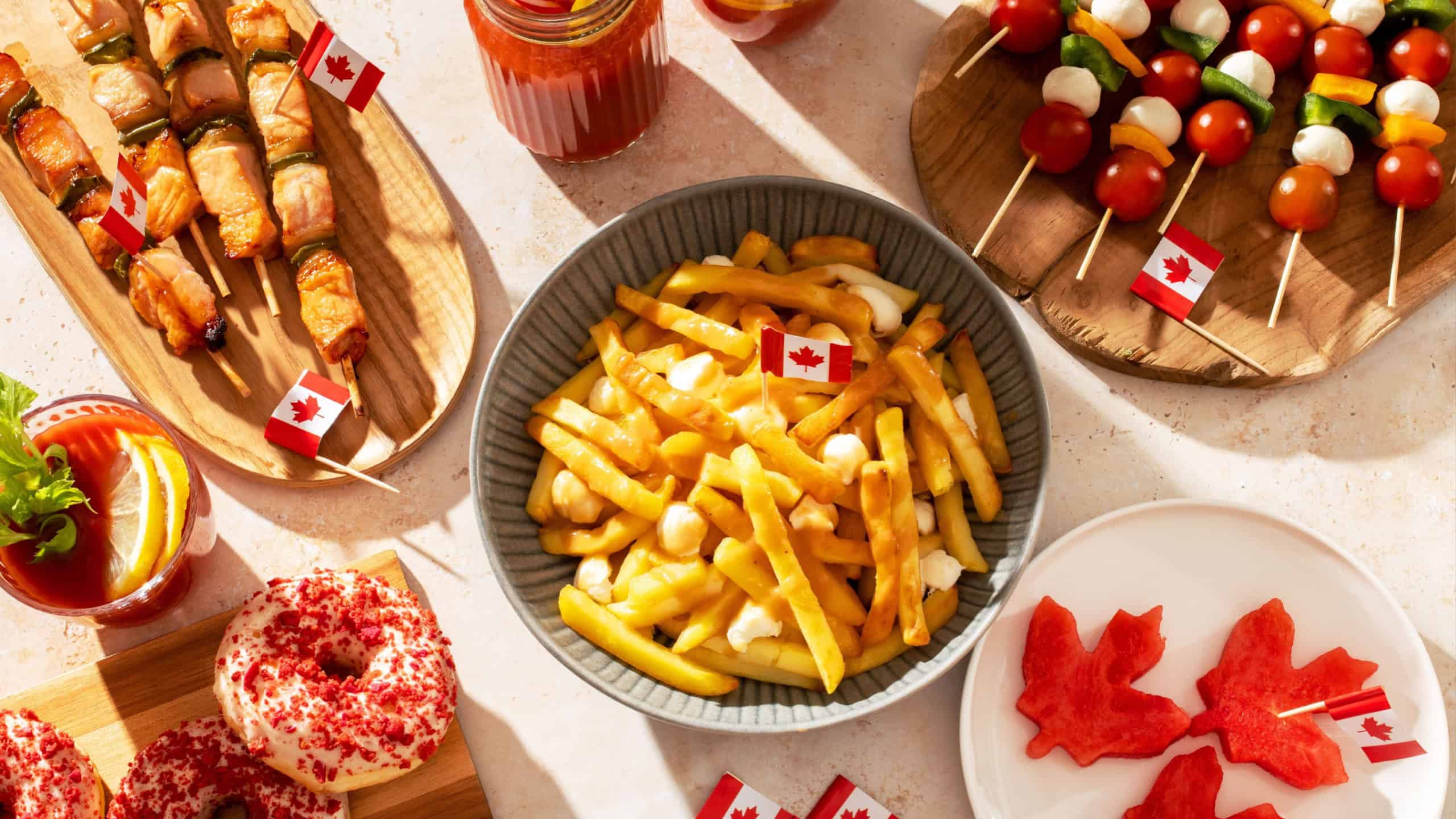 Top view of Canada Day party food with poutine, hot dogs, fruit, skewers, and donuts decorated with Canadian flags