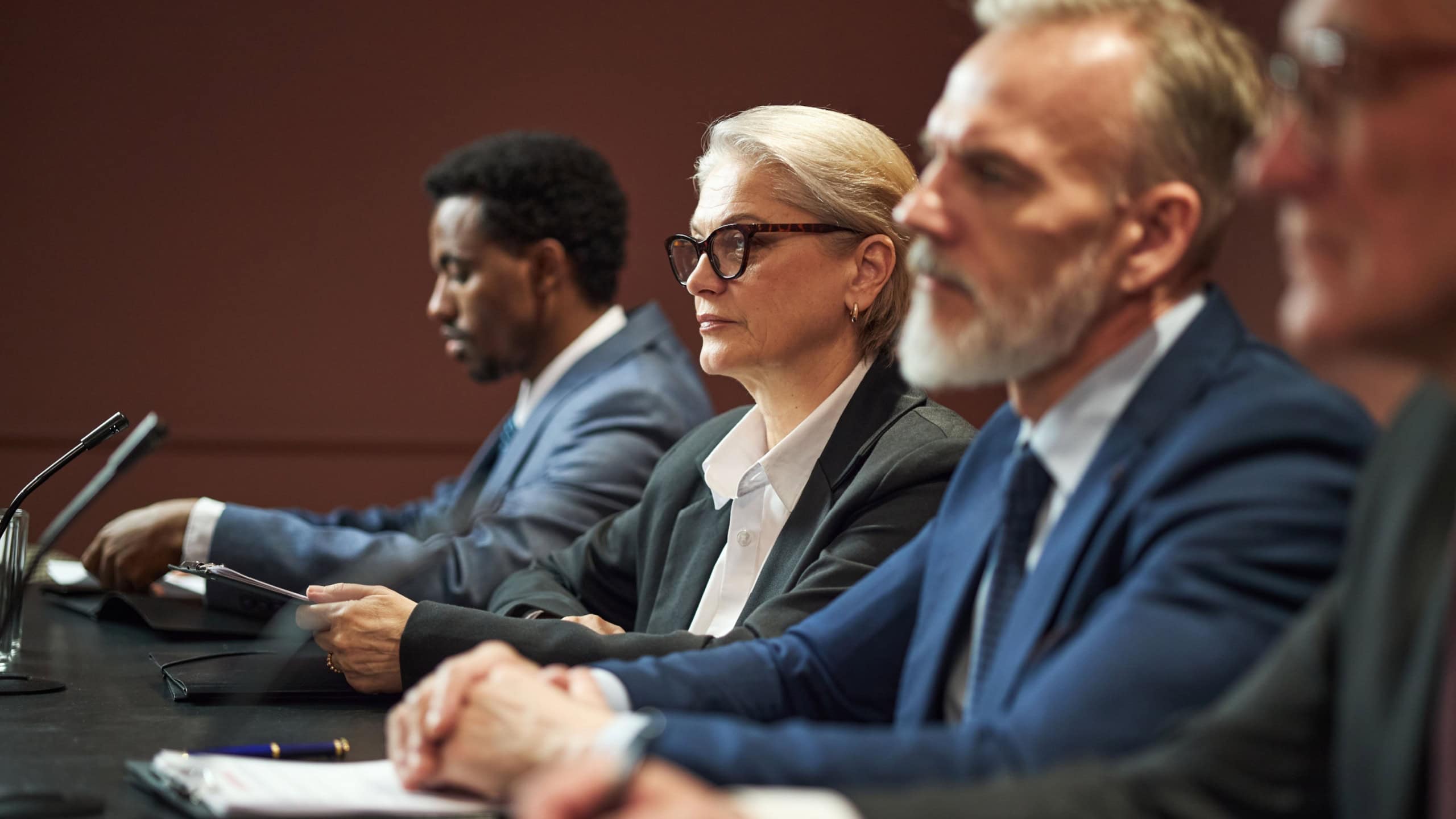Middle aged Caucasian woman and middle aged Caucasian man sitting at conference table with Black man and another man, participating in political meeting, microphones visible