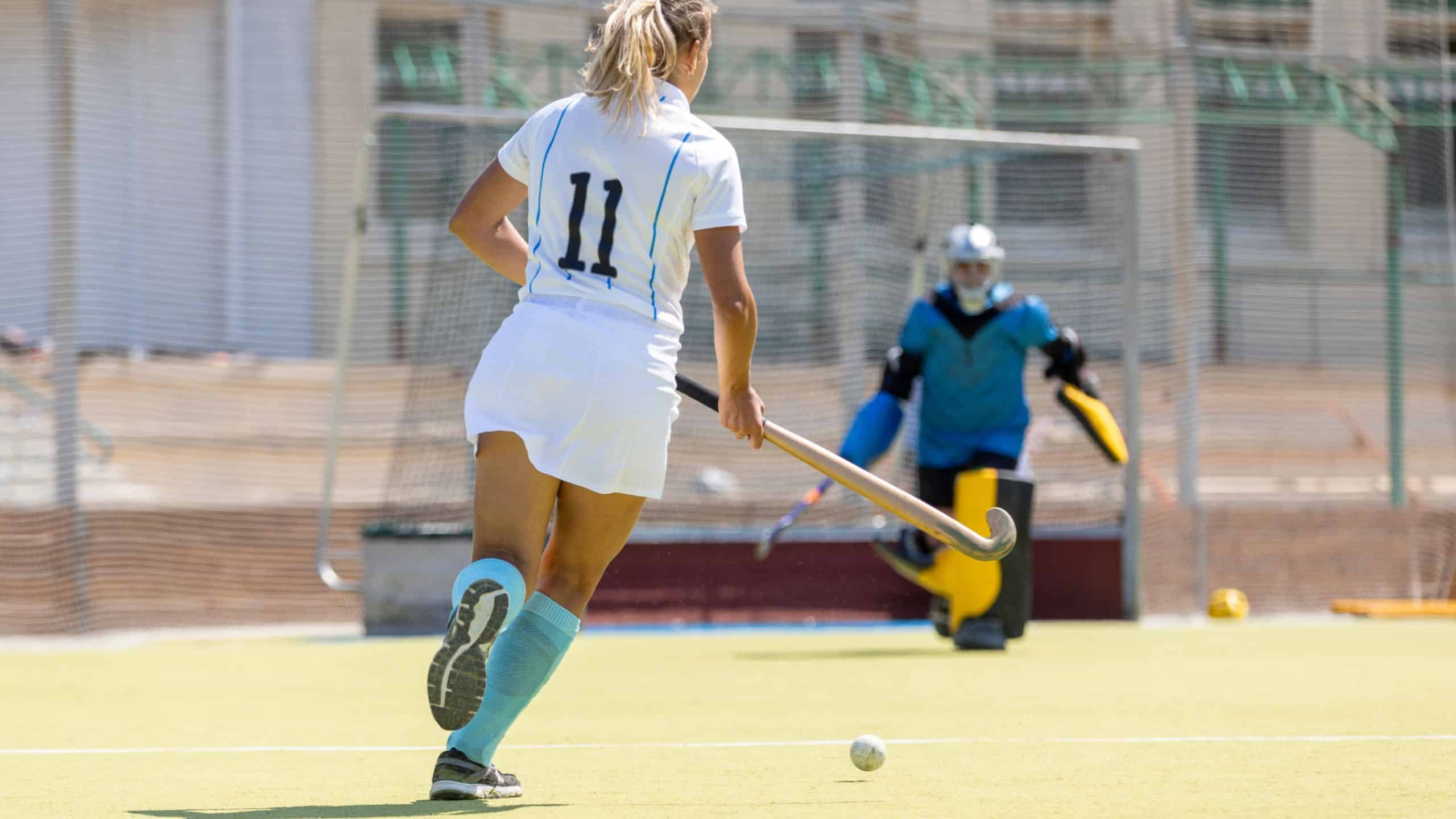 A player in white prepares to shoot at the goal during a field hockey match on a sunny day at the sports facility.
