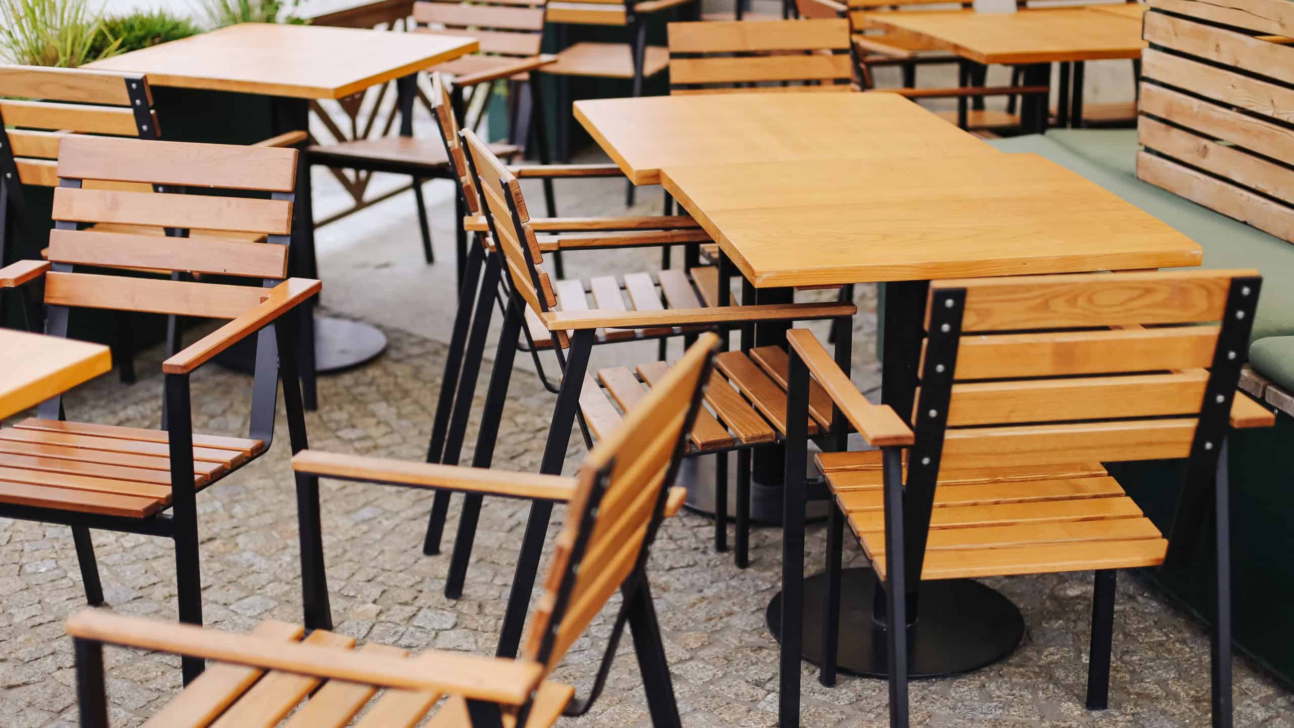 Wooden tables and chairs at outdoor cafe terrace. Empty restaurant furniture arranged on cobblestone pavement. Modern design with wooden seats and metal frames. Cozy atmosphere of urban dining area.