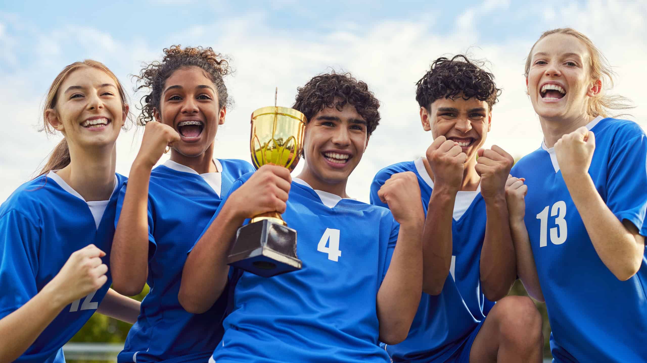 Excited school team celebrating with a golden trophy after a match. Smiling young players cheering and holding a championship cup. Joyful group of teammates showing pride and happiness.