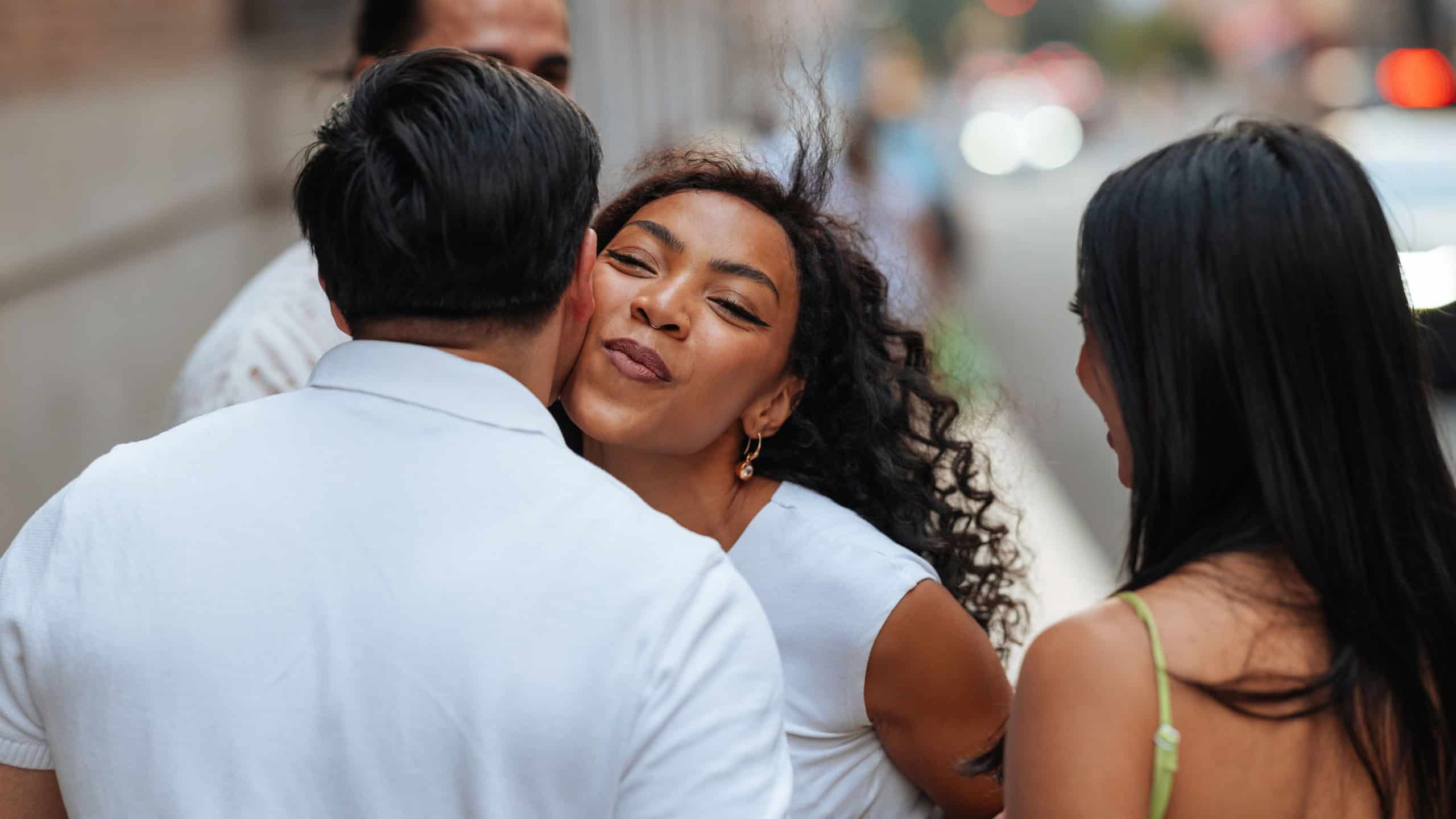 Tourists joyfully greeting each other on a bustling city street, with a woman affectionately kissing a man on the cheek, capturing a moment of connection