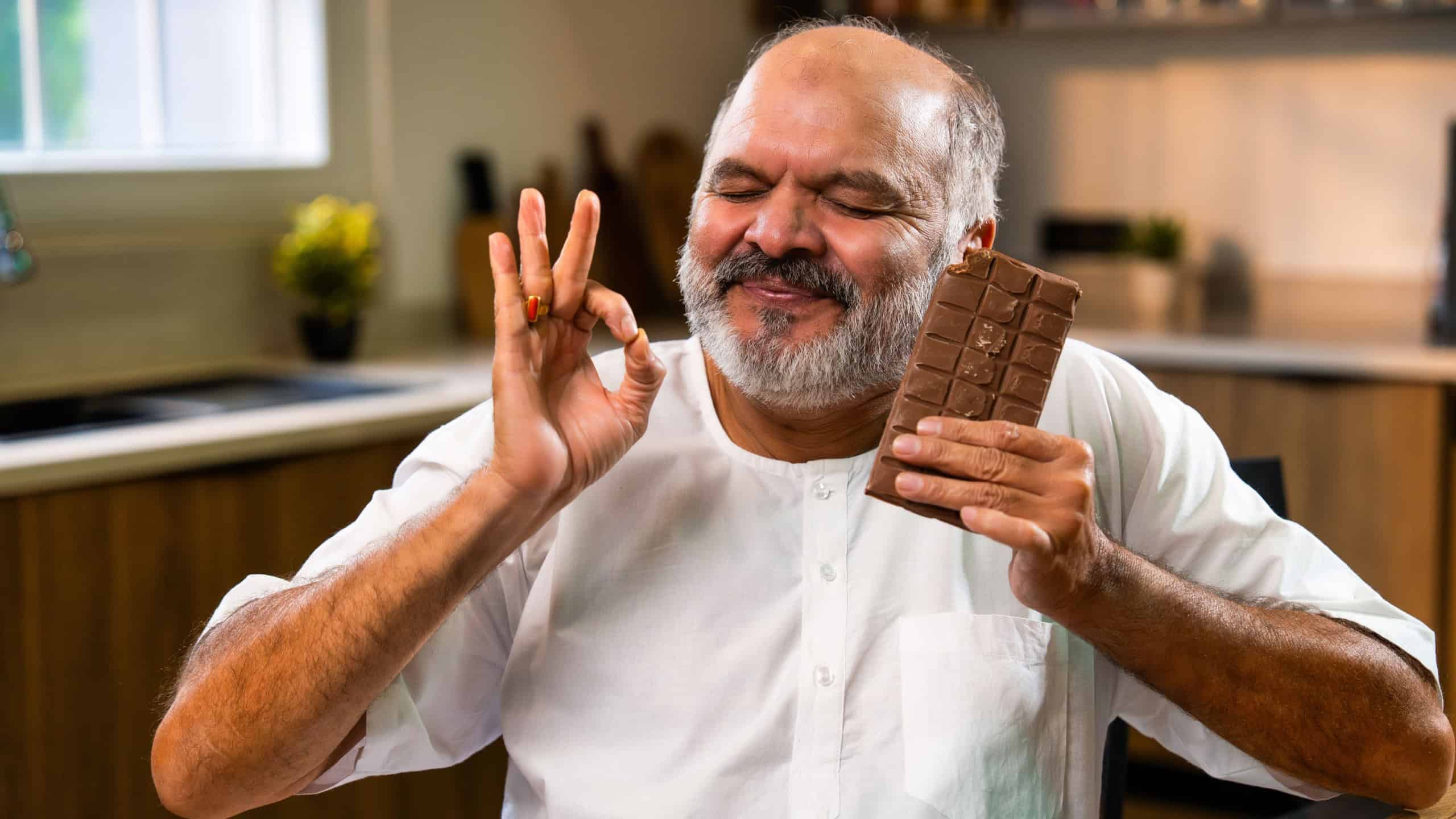 Senior man secretly eating chocolate brick in modern kitchen, Asian elderly foodie sneaking thick chocolate slab with mischievous expression, enjoying sweet treat alone at home