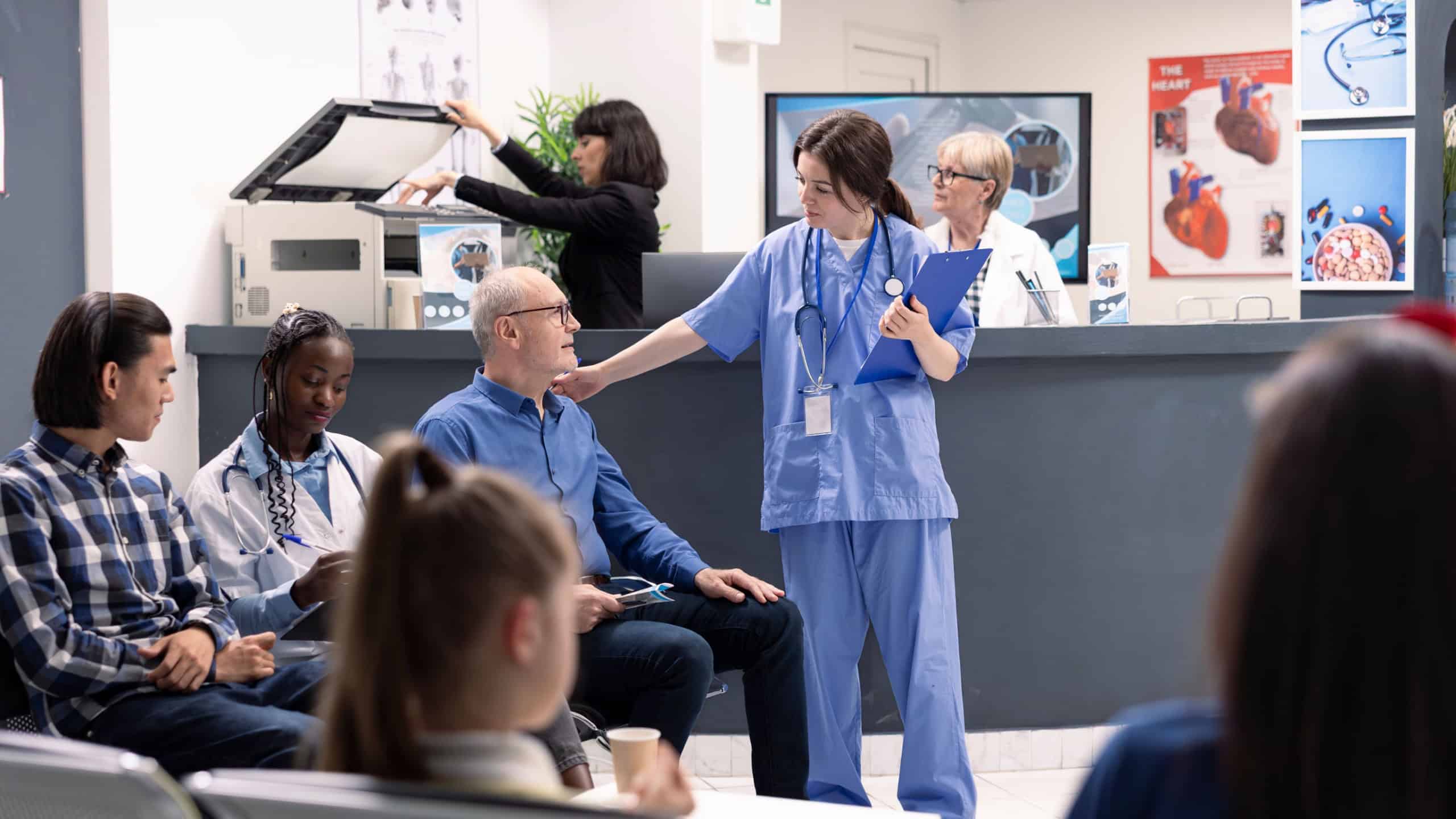 Young nurse in blue scrubs speaking with elderly man seated in hospital waiting room. Healthcare professional offering assistance with care and comfort during medical appointment.