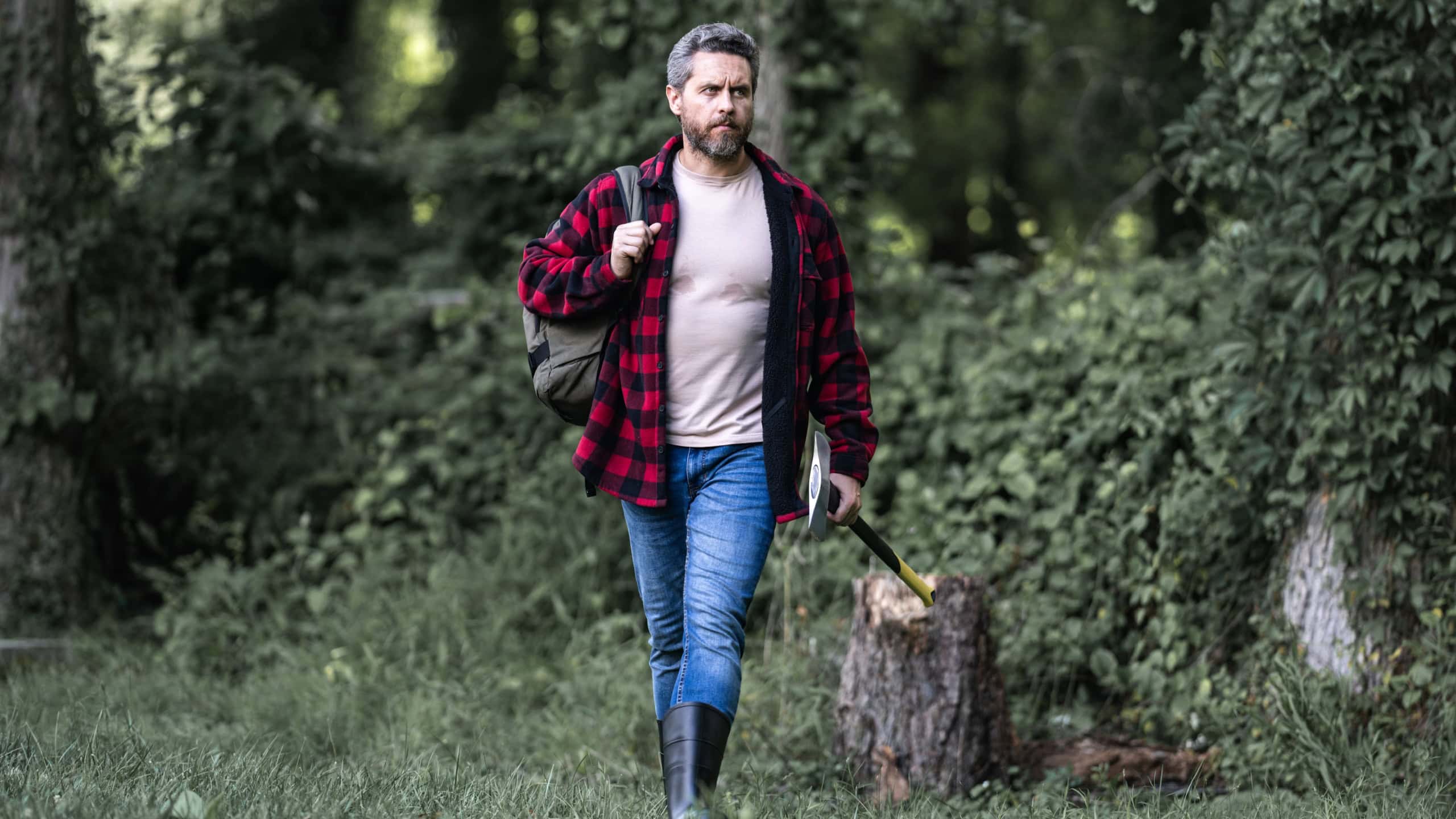 The Lumberjack holds axe with serious face on the forest. A lumberjack with a large ax examines the tree before felling. Serious lumberjack. Confident young bearded man holding a big axe outdoor.