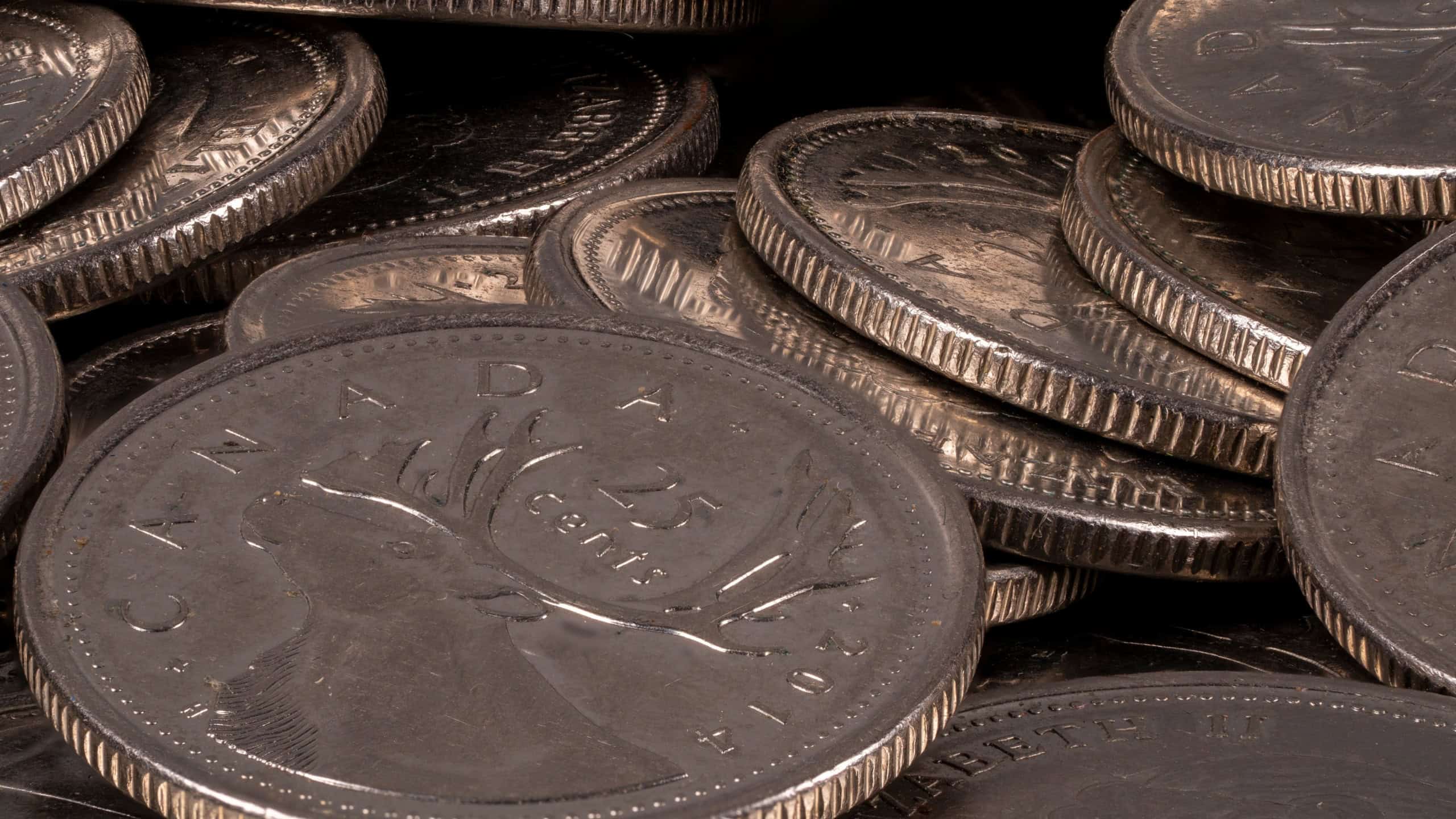 Many Canadian coins are scattered on a surface. The coins display various designs and are stacked in a disorganized manner. Light reflects off their surfaces.