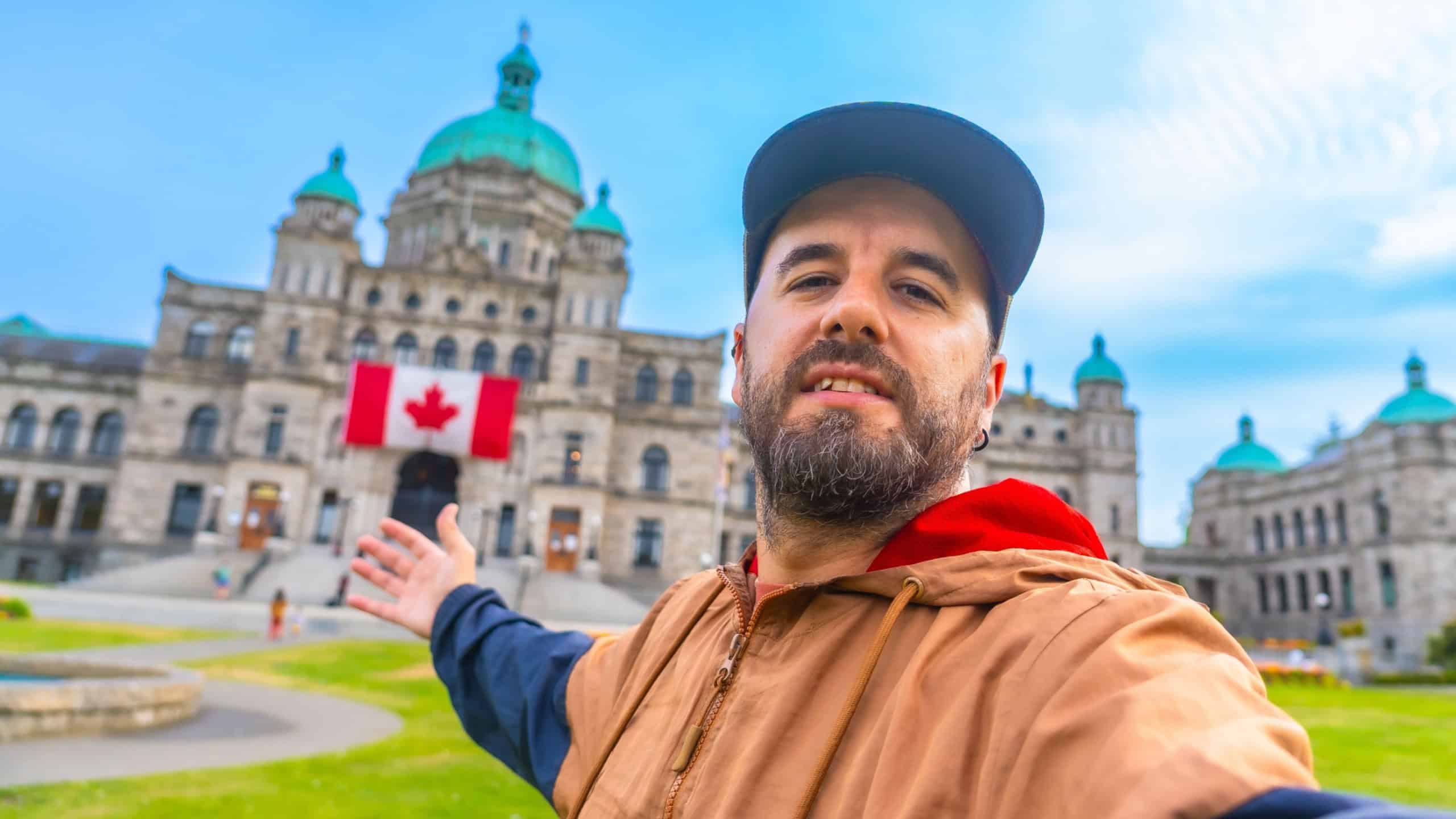 Happy bearded tourist taking a selfie with outstretched arms in front of the parliament buildings with a canadian flag hanging from the facade in victoria, british columbia, canada