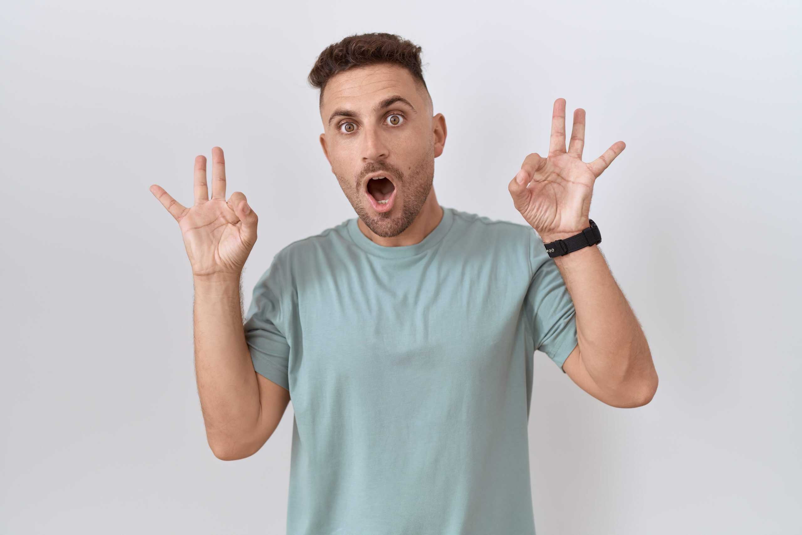 Hispanic man with beard standing over white background looking surprised and shocked doing ok approval symbol with fingers. crazy expression
