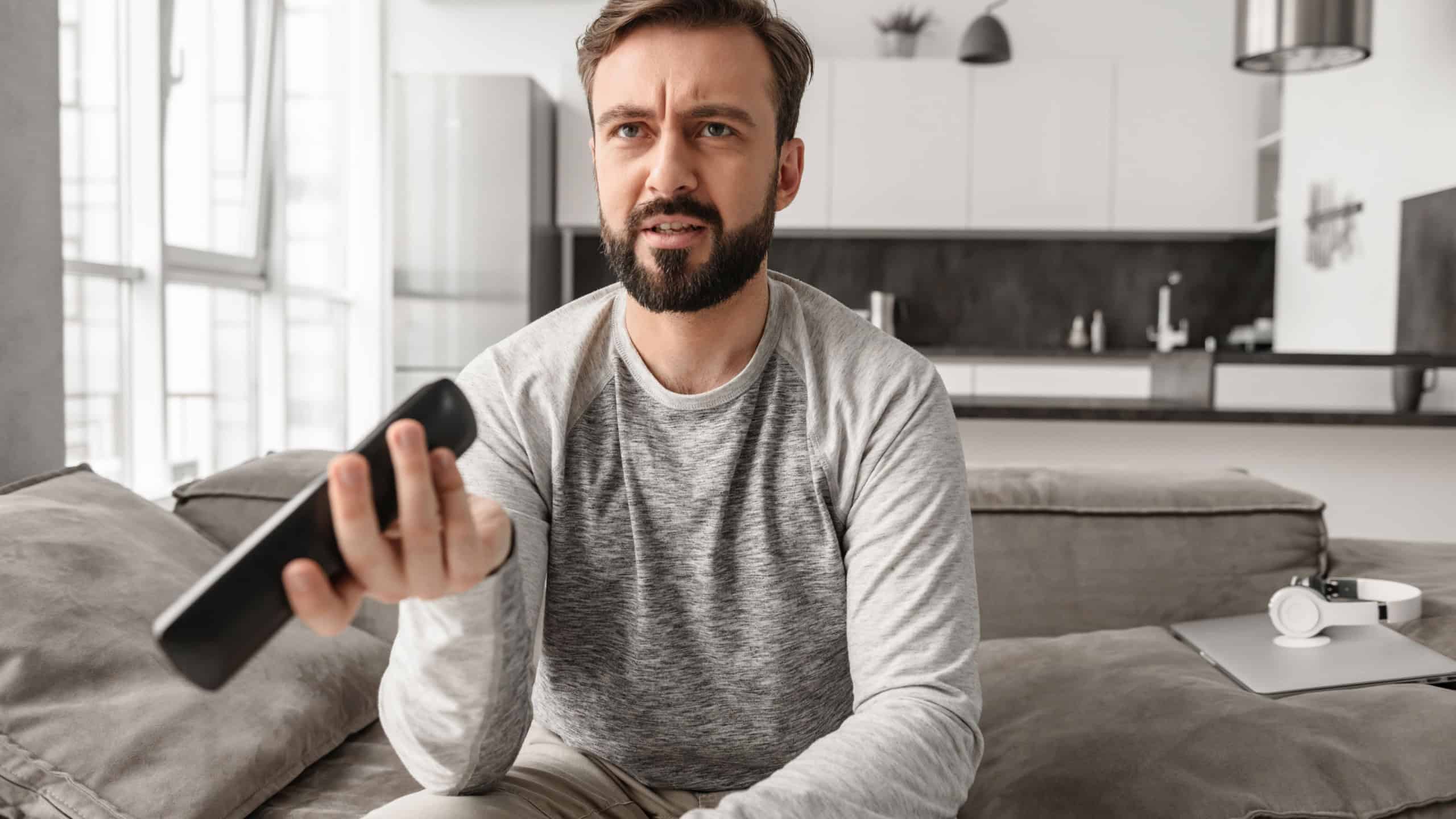 Portrait of a disappointed young man holding TV remote control while sitting on a couch at home