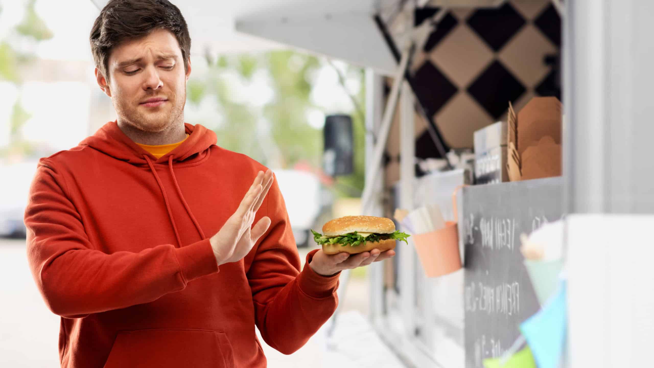 Fast food, unhealthy eating and people concept - young man in red hoodie refusing from hamburger over food truck on street background