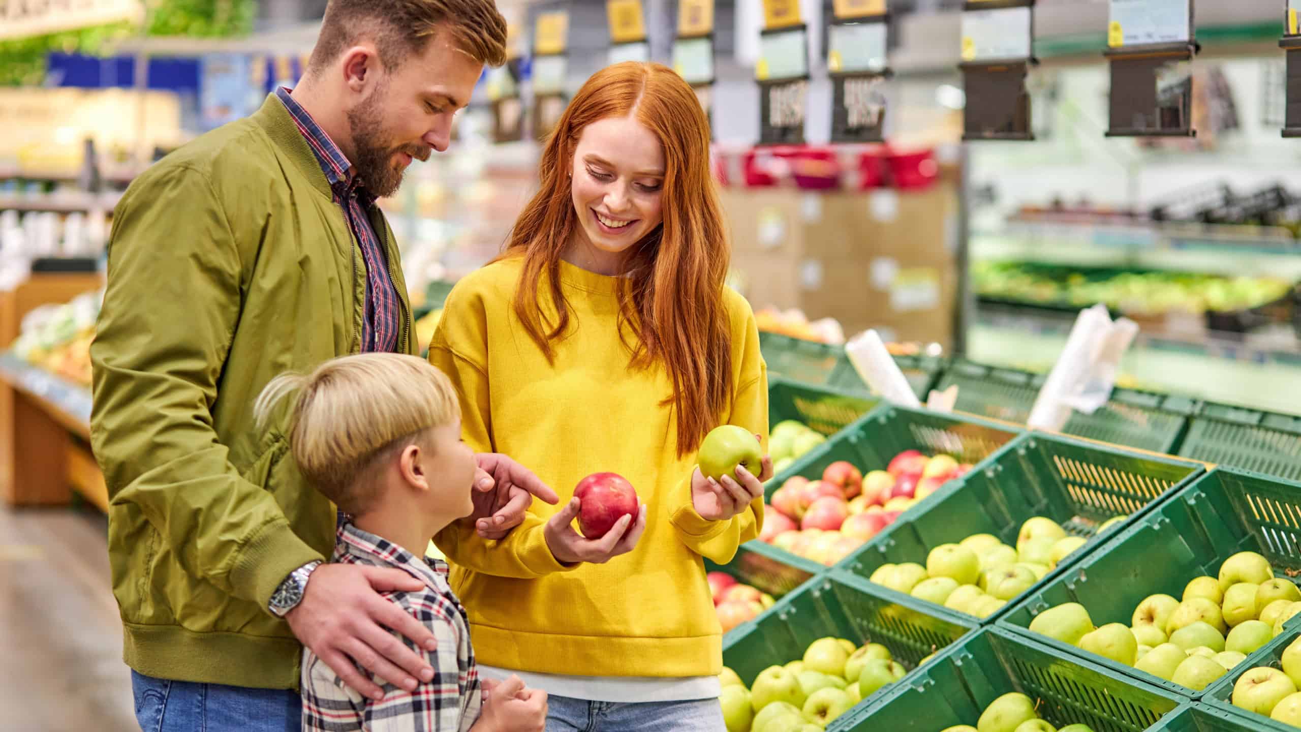 Husband and wife with a kid buy fruits, apples. family of three choosing fresh apple in fruits department of supermarket or market