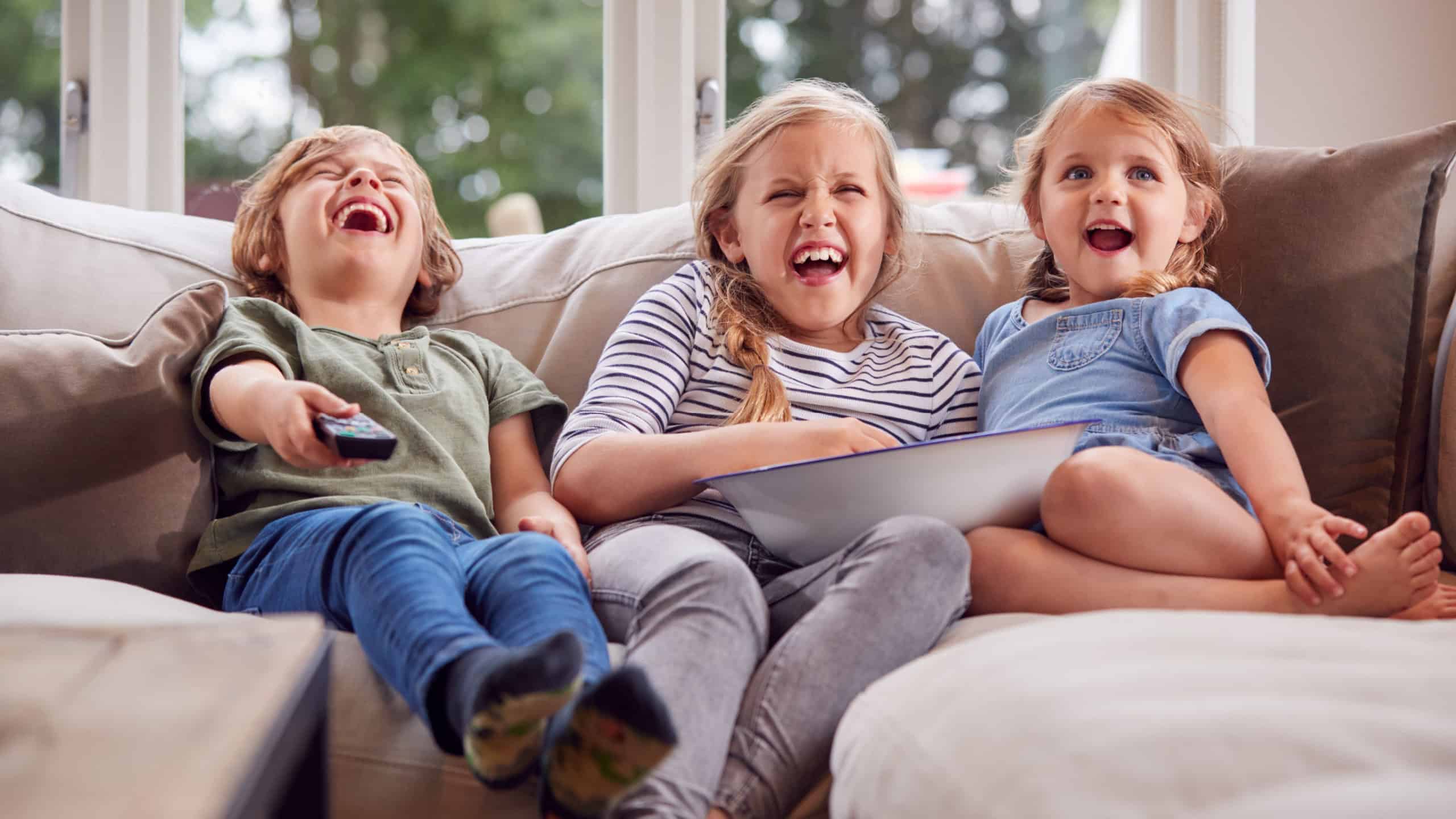 Three Children Sitting On Sofa At Home Laughing And Watching TV With Popcorn