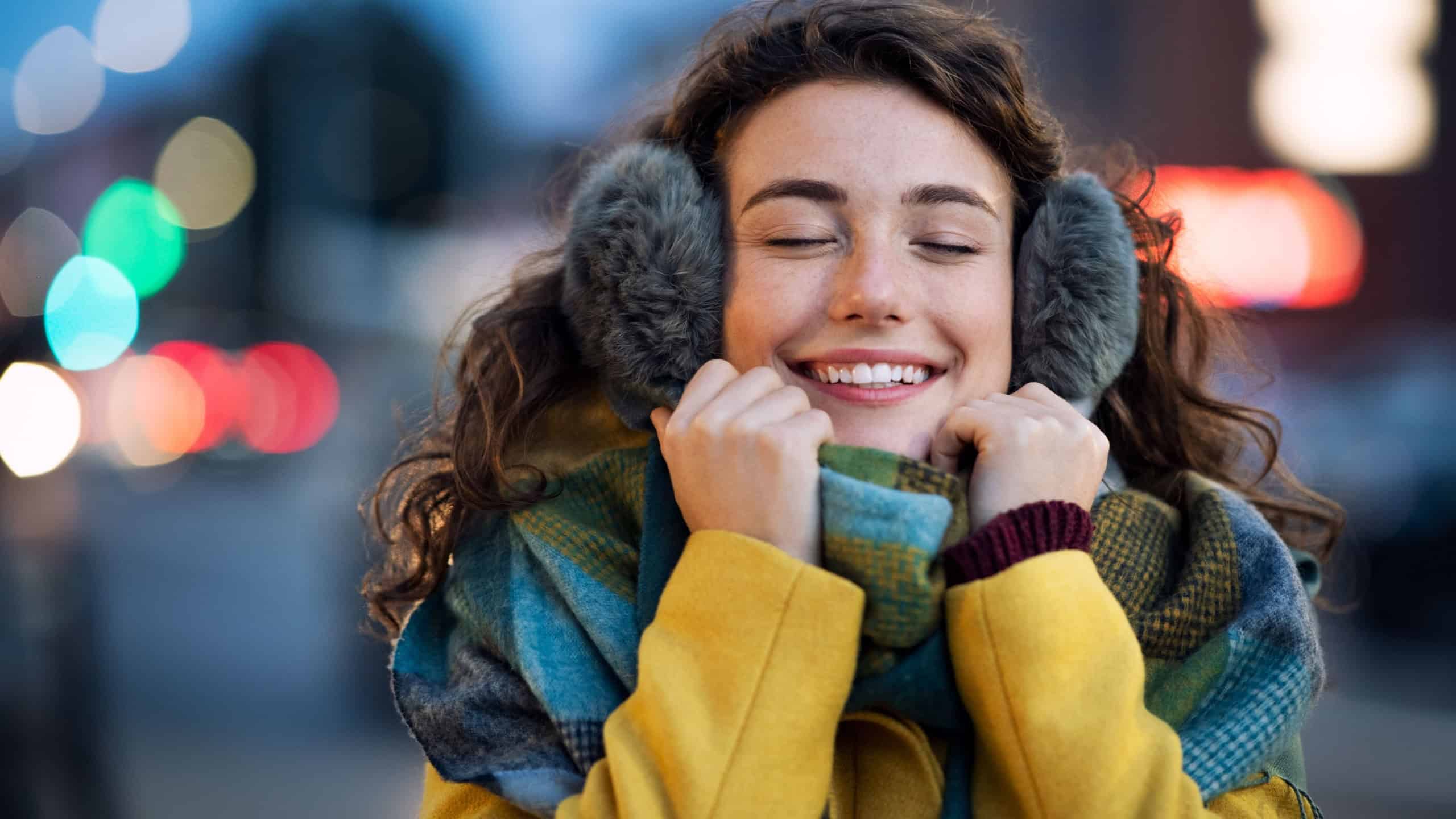 Young happy woman wearing winter jacket at dusk with closed eyes. Pretty girl with warm scarf and yellow coat standing outdoors on winter evening with urban background. Girl with ear muffs in dusk.