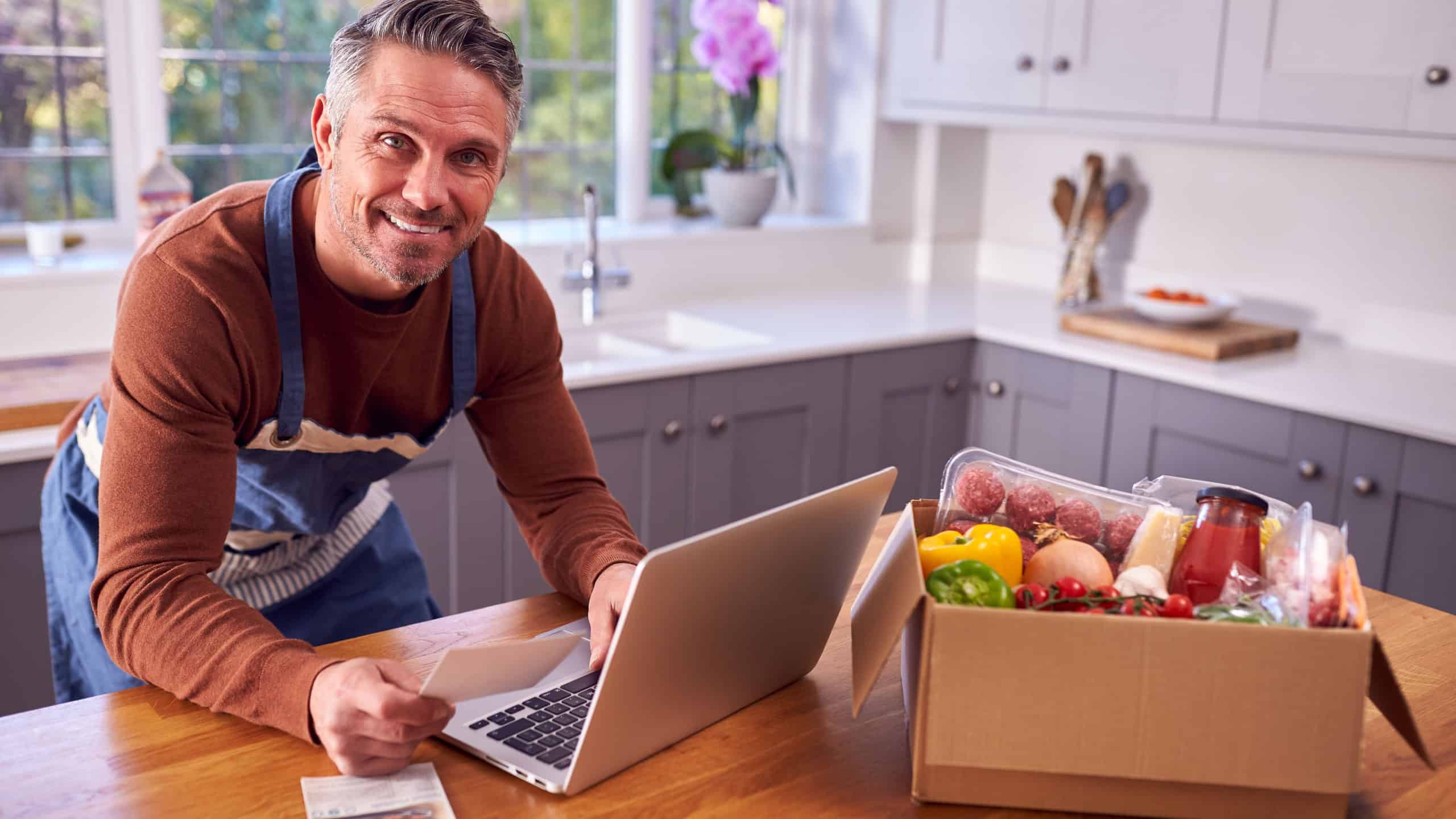 Mature Man In Kitchen With Laptop To Find Recipe For Online Meal Food Kit Delivered To Home